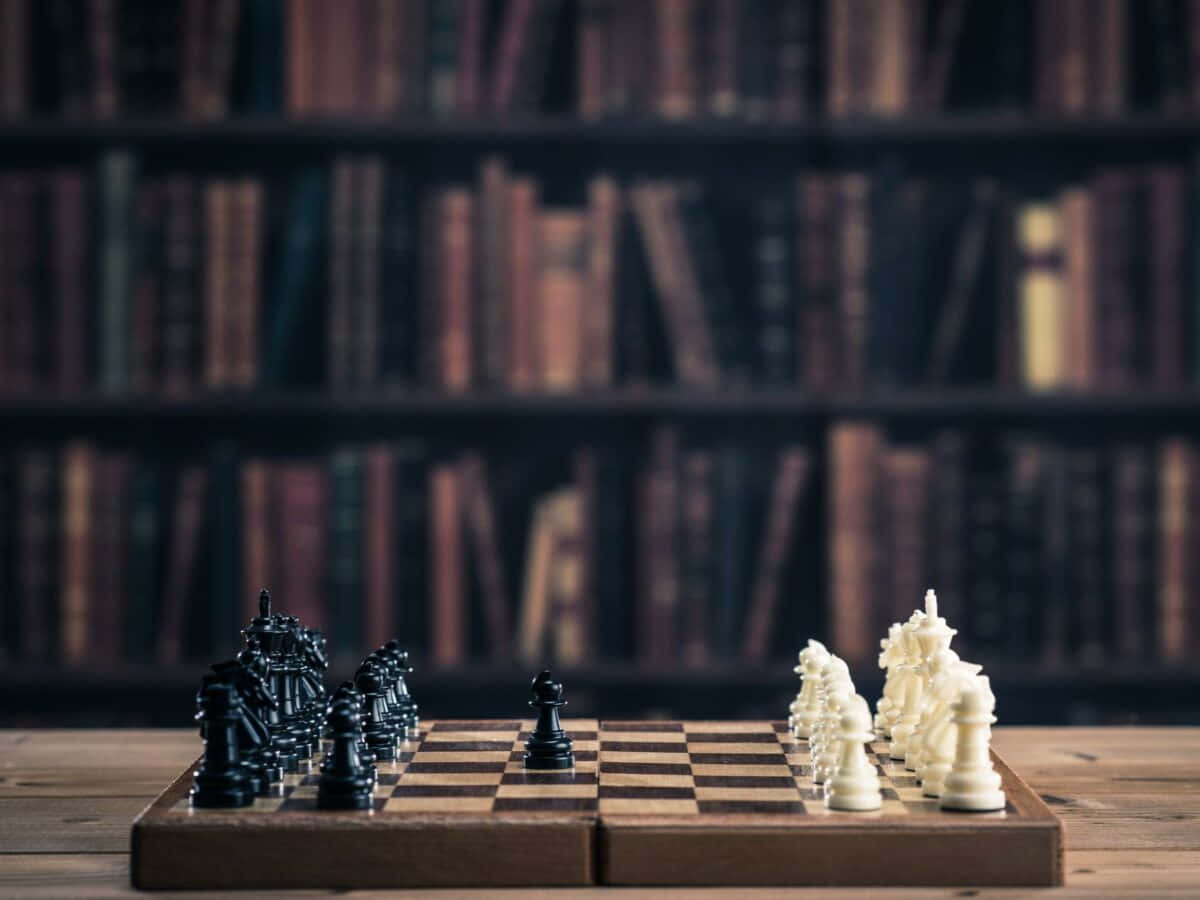 Chessboard 桌布 壁紙 — 一個木製棋盤上擺放著黑白棋子，準備對弈，放在一張桌子上，背景是模糊的書架，上面擺滿了書。
