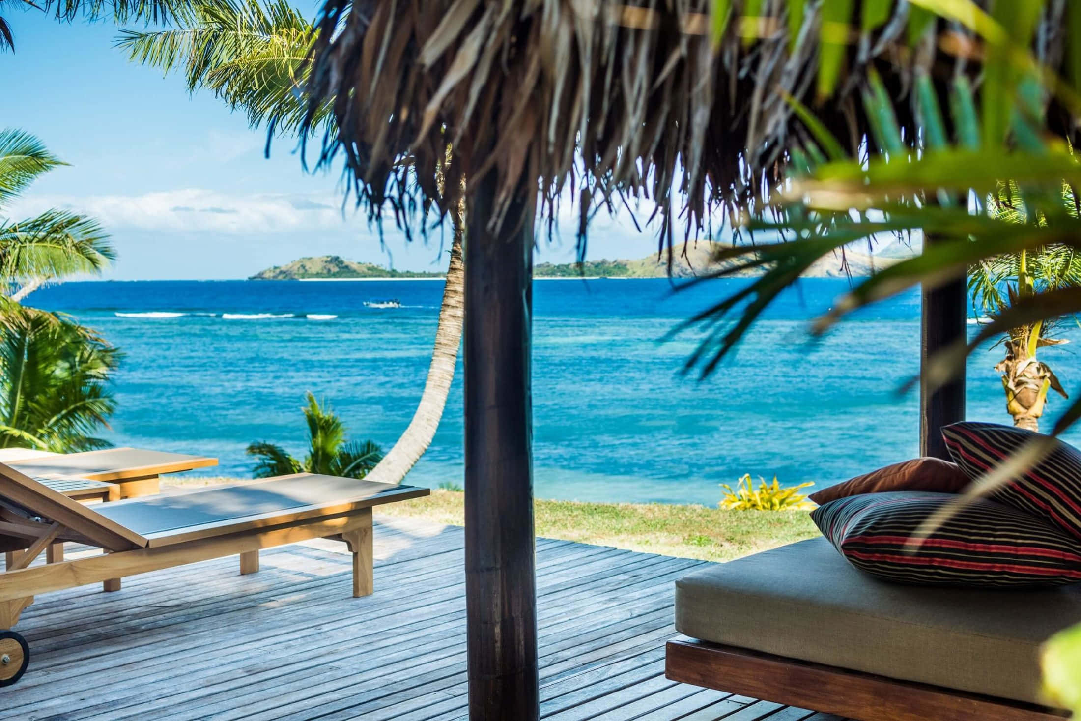 Fiji Beach 桌布 壁紙 — 一個木製甲板，茅草屋頂下設有躺椅，俯瞰著湛藍的海洋和遠處的島嶼，天空明亮湛藍。
