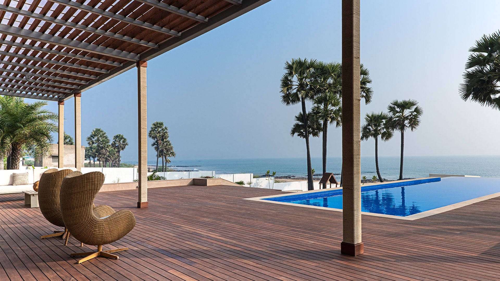Beach House 桌布 壁紙 — 一個木製甲板配以柳條椅，俯瞰著藍色無邊際泳池和棕櫚樹環繞的海洋，天空晴朗。
