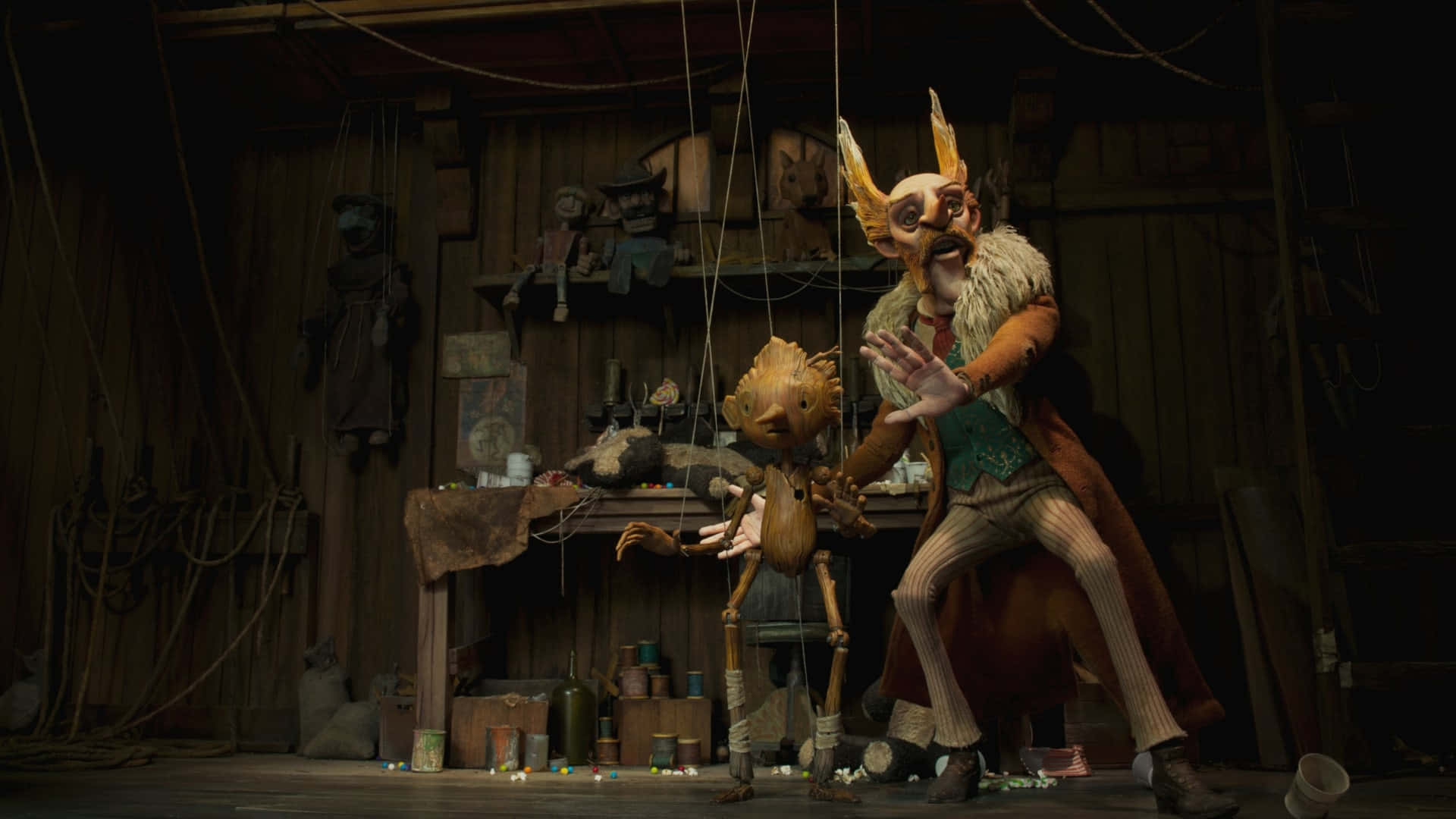 Guillermo Del Toro's Pinocchio 桌布 壁紙 — 一個木製的操偶師，頭上有角，留著鬍子，在一個雜亂昏暗、佈滿繩索和工具的工作室裡操控一個小木偶。