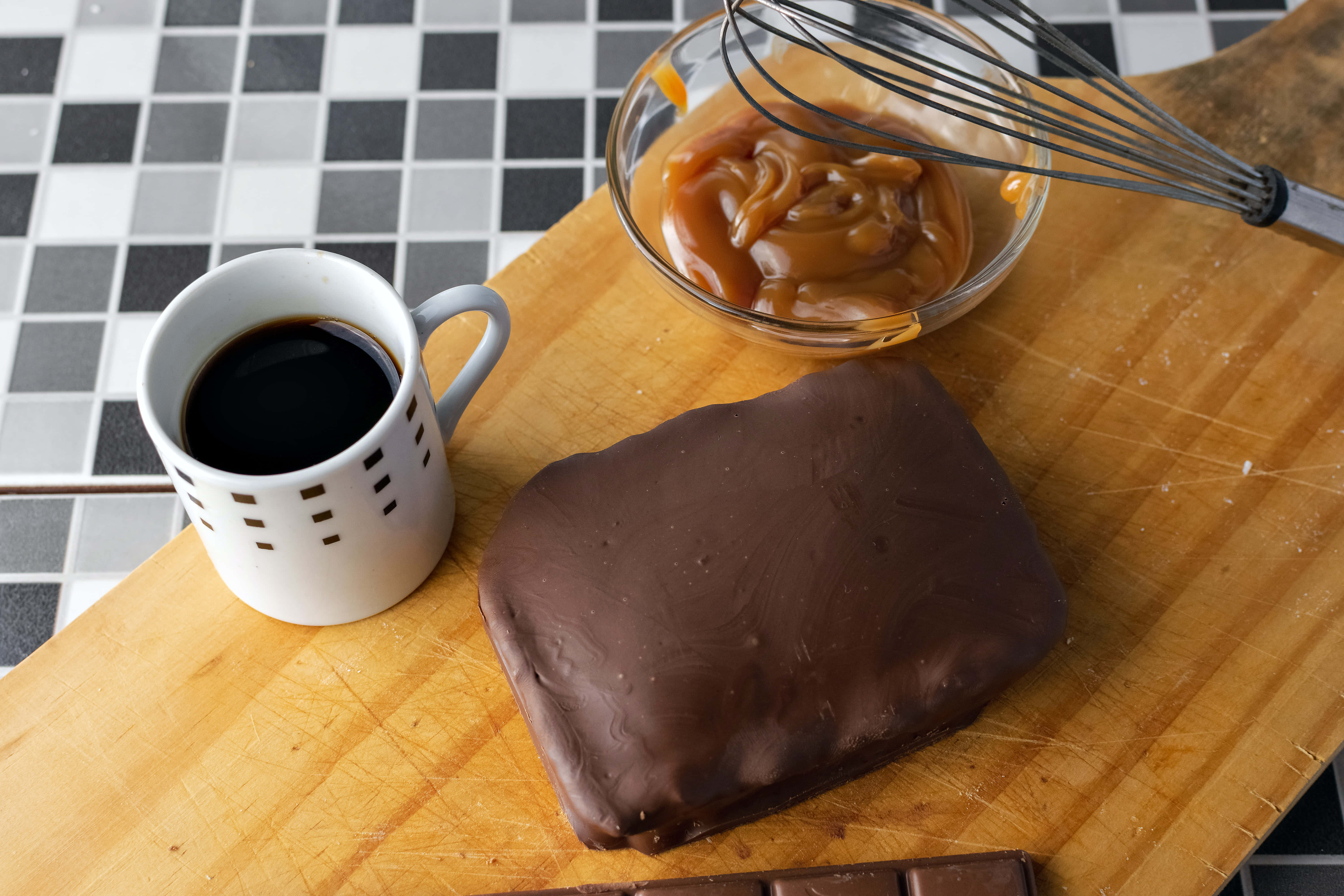 Caramel 桌布 壁紙 — 一個木製砧板上放著一塊巧克力焦糖糖果、一碗焦糖醬和一杯黑咖啡，置於格子瓷磚檯面上。