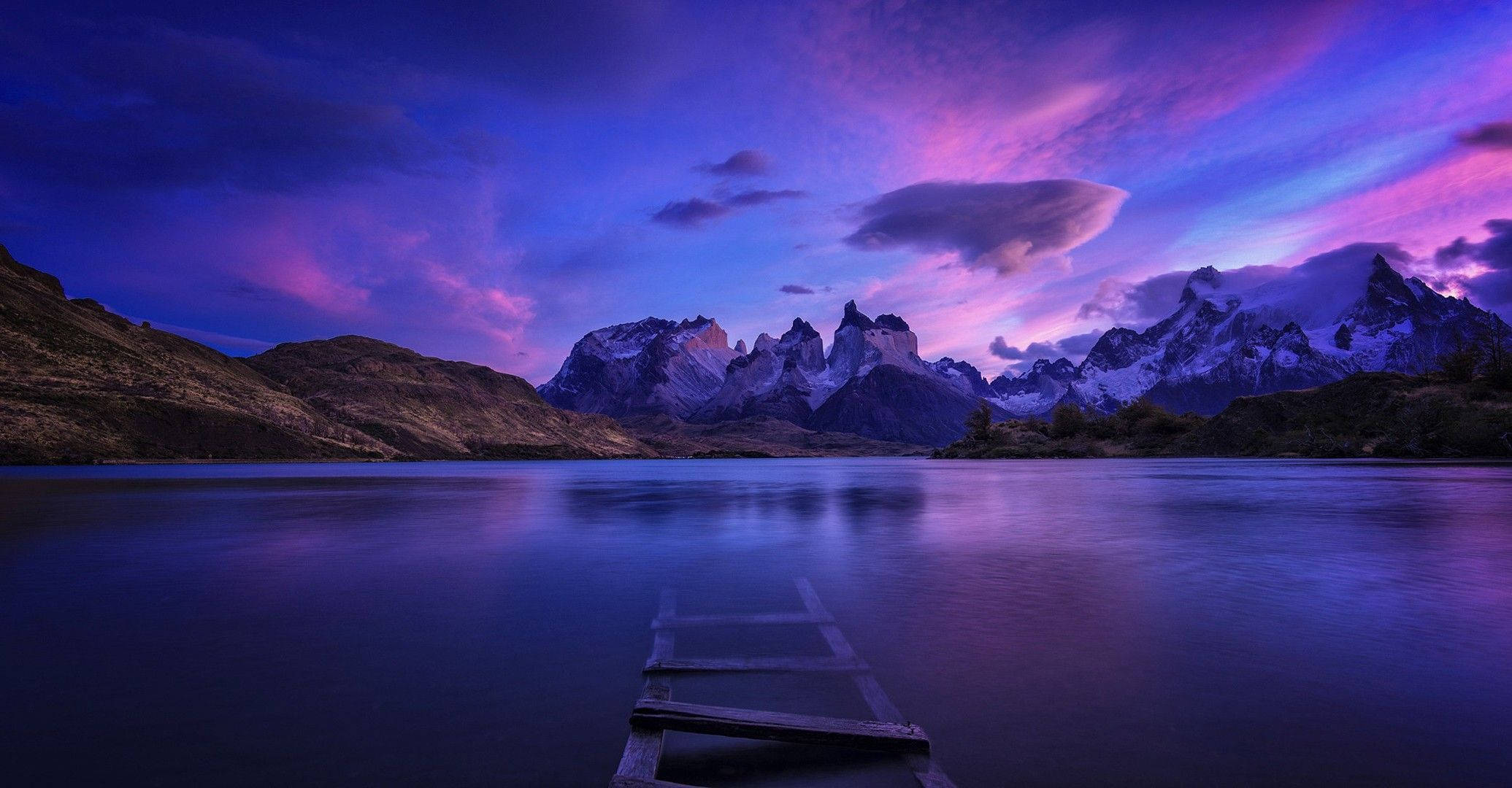 Patagonia 桌布 壁紙 — 一個木製碼頭延伸到平靜的湖面，湖面倒映著白雪覆蓋的山脈，天空呈現出鮮豔的紫色和粉紅色暮色。
