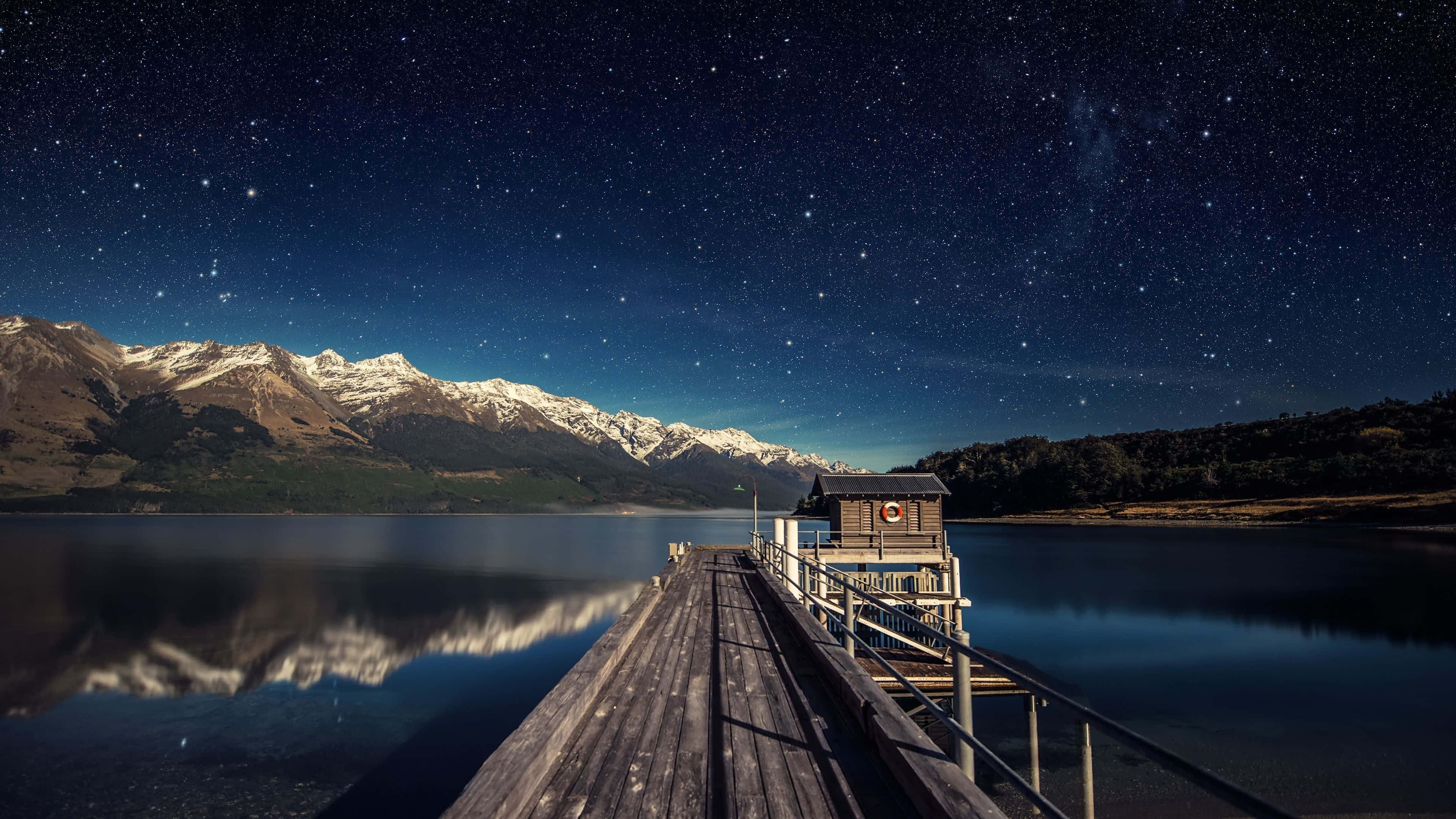 4K Computer 桌布 壁紙 — 一個木製碼頭延伸到繁星點點的夜湖中，積雪的山脈倒映在平靜的水面上。