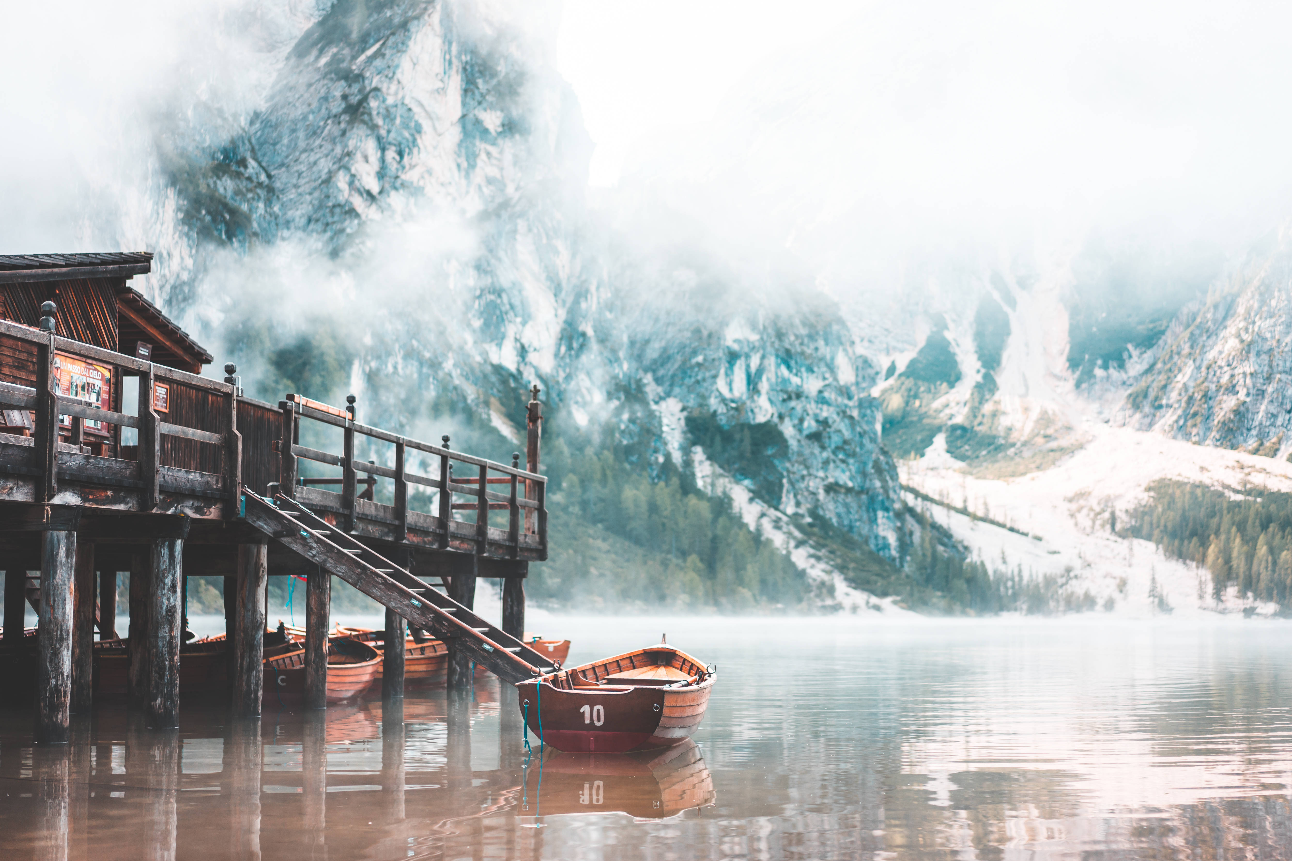 HD Scenery 桌布 壁紙 — 一個木製碼頭延伸到霧氣瀰漫的湖面上，附近停泊著一艘小船，周圍是白雪覆蓋的高聳山脈。