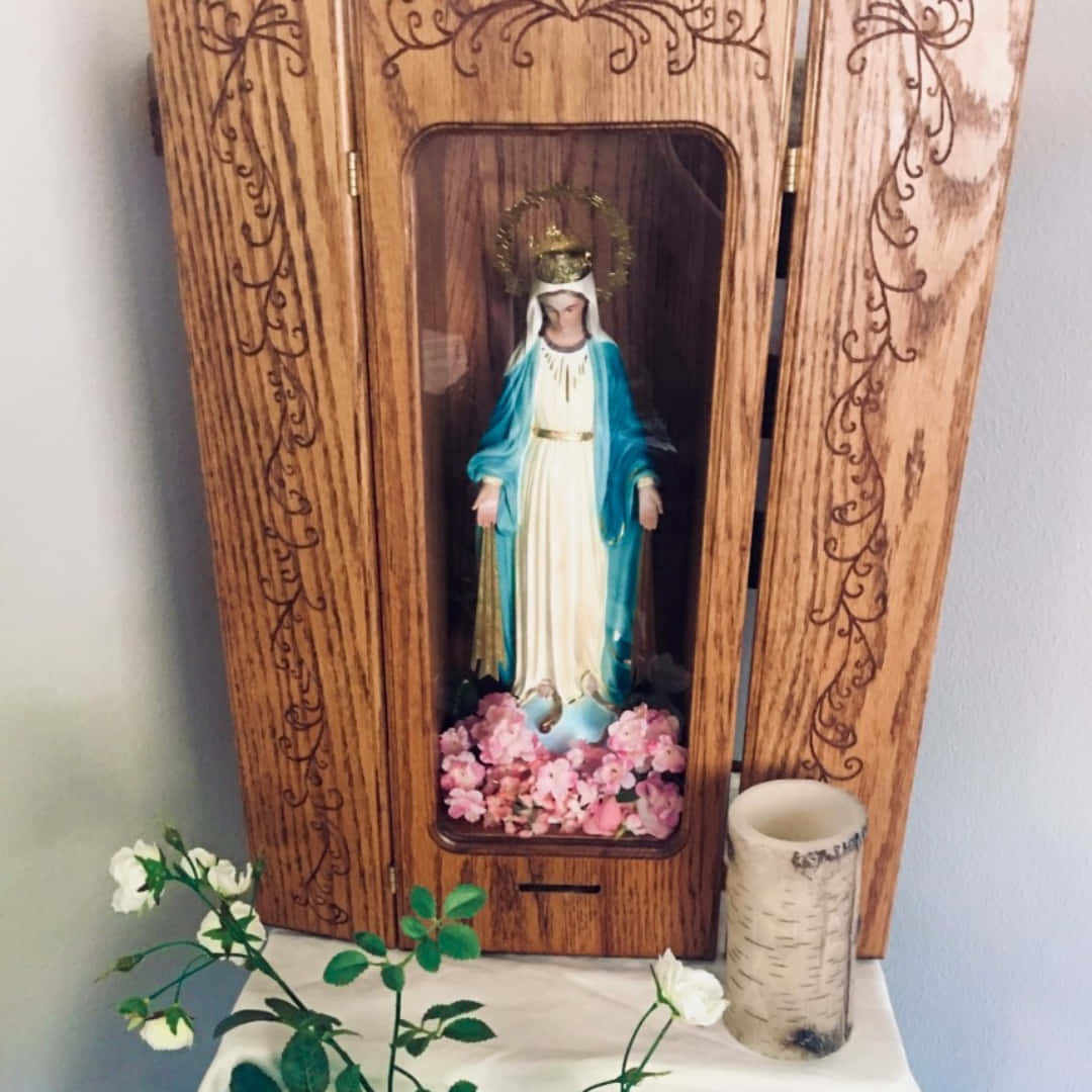 Mother Mary 桌布 壁紙 — 一個木製神龕展示了一尊聖母瑪利亞的雕像，她穿著藍色和白色的長袍，周圍有粉紅色的花和一支蠟燭。