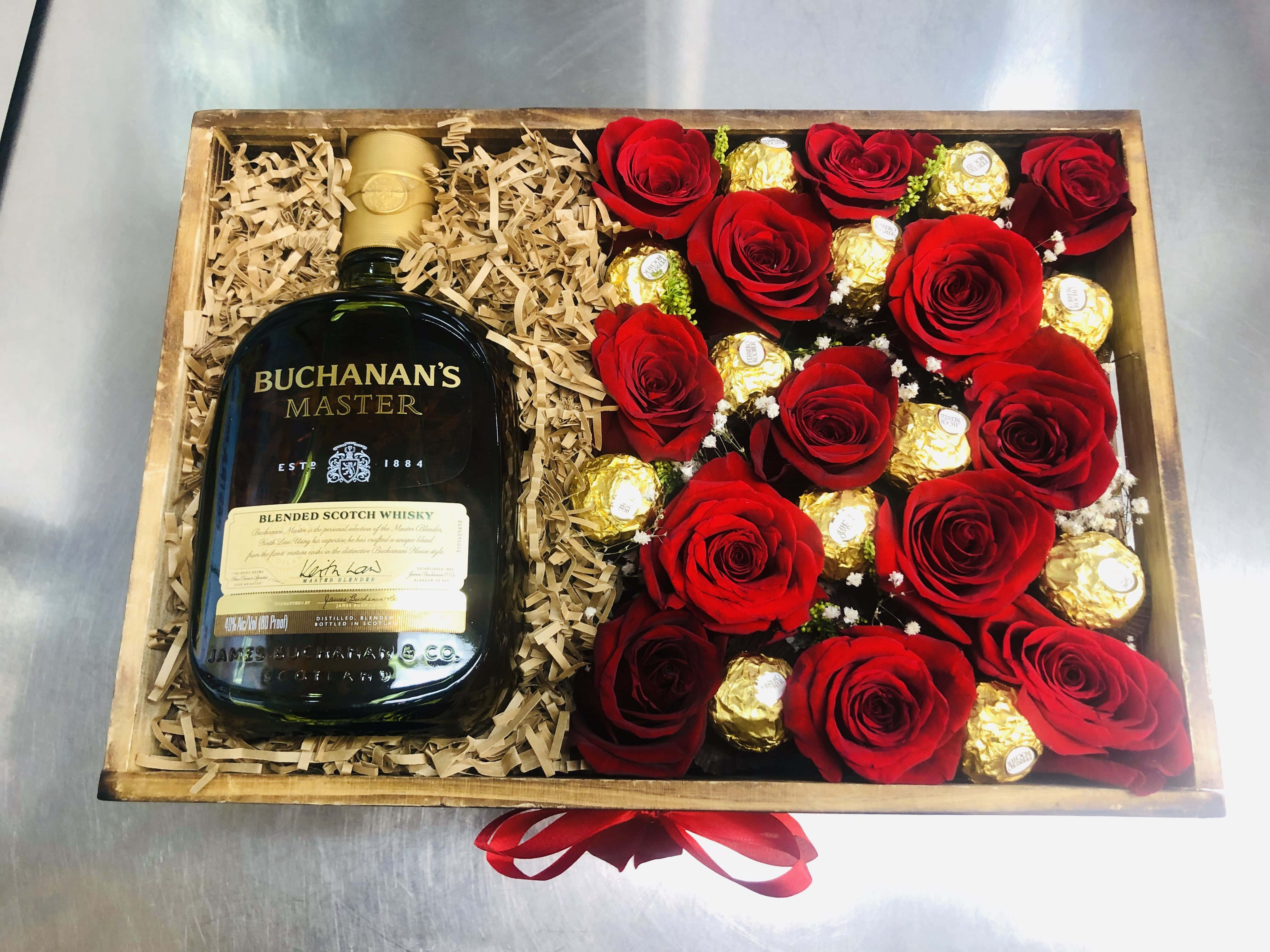 Gift 桌布 壁紙 — 一個木製禮品盒內有一瓶Buchanan's Master調和蘇格蘭威士忌，放在木屑中，旁邊是鮮豔的紅玫瑰和金色包裝的巧克力。