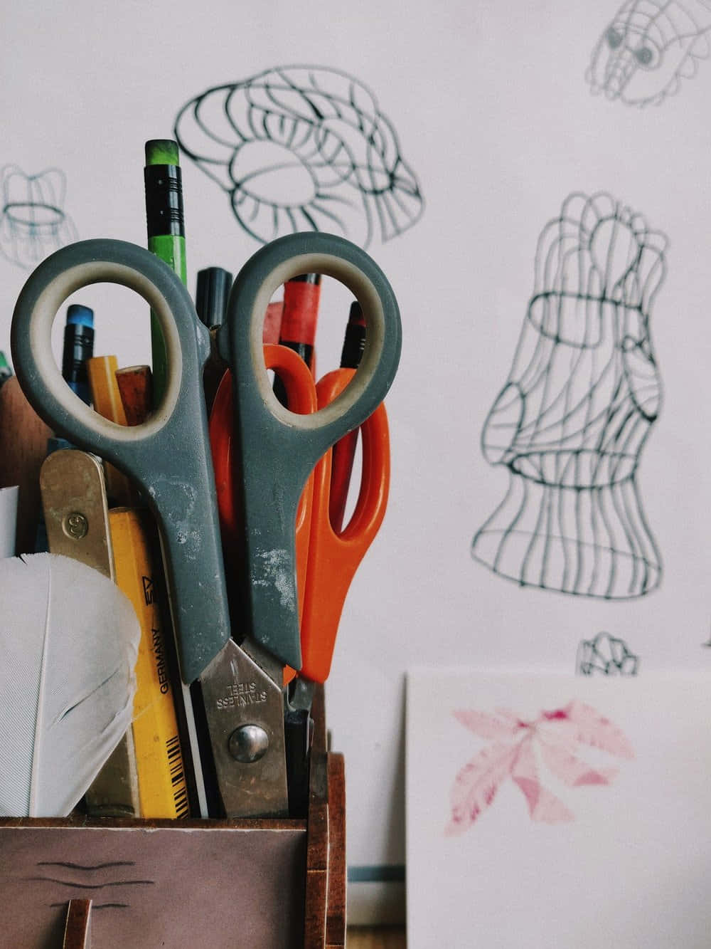 Clothing Design 桌布 壁紙 — 一個木製筆筒裝滿了美術用品，包括剪刀、鉛筆和一根羽毛，背景是一幅素描畫牆。