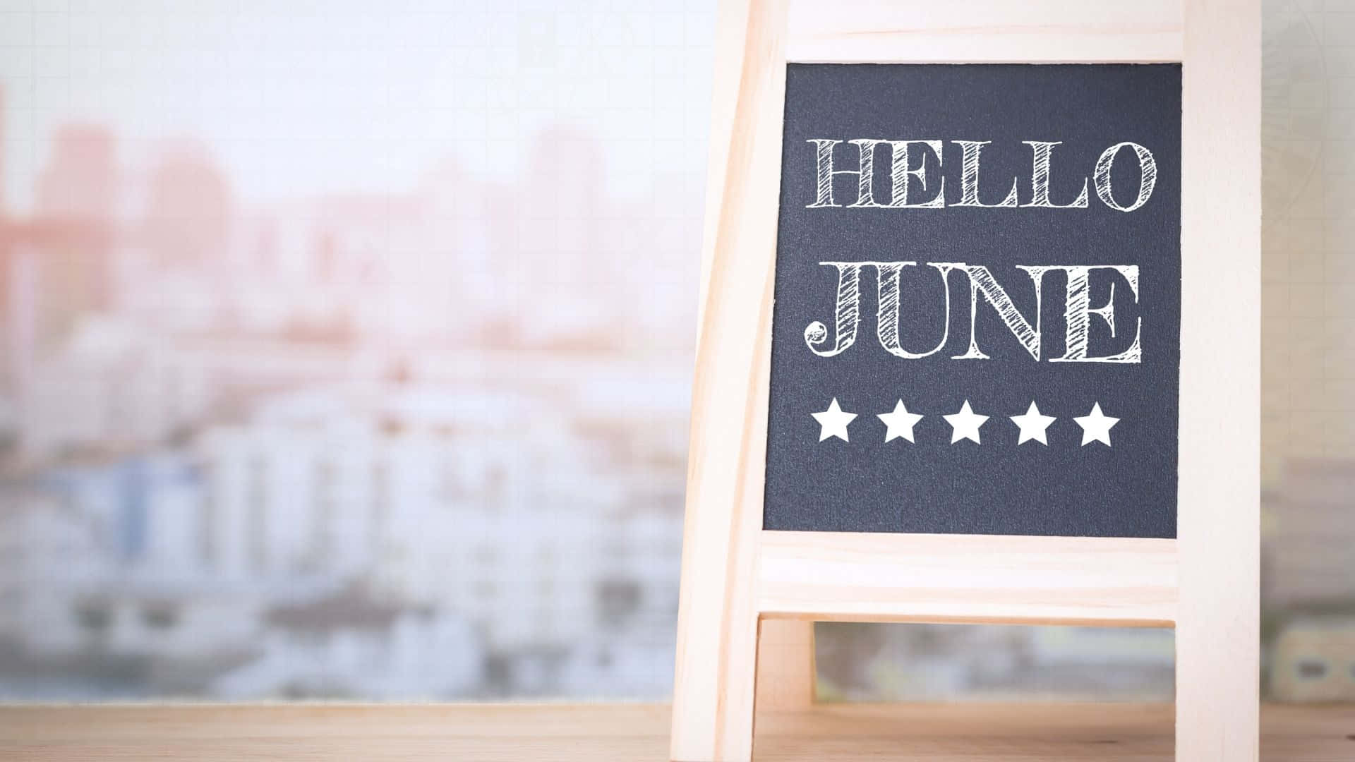 June 桌布 壁紙 — 一個木製黑板標牌，上面寫著「HELLO JUNE」，並有五顆星星，放在一張桌子上，背景是模糊的城市景觀。