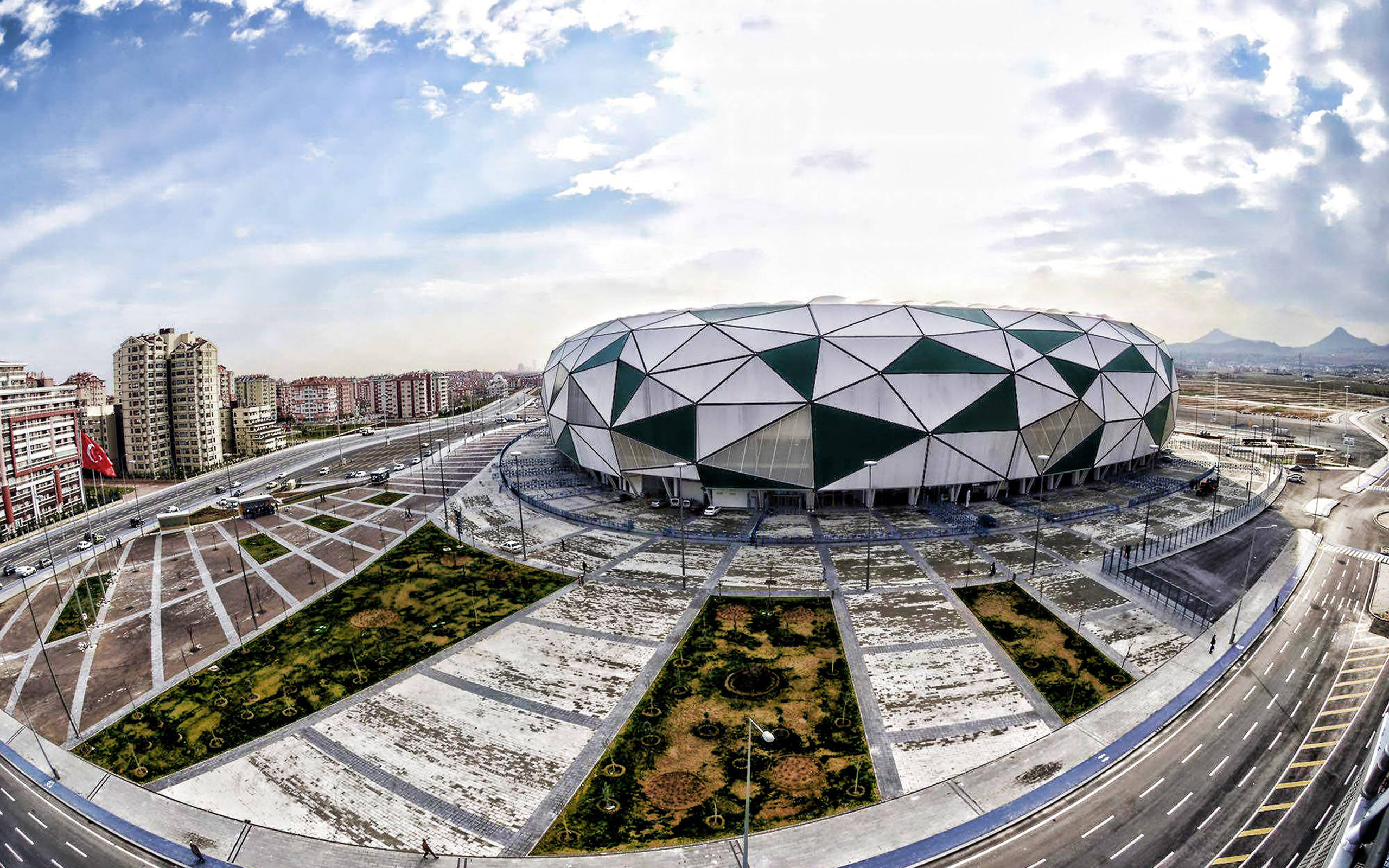Konya 桌布 壁紙 — 一個未來主義的圓頂體育場，帶有幾何綠色和白色面板，在部分多雲的天空下突出地矗立在城市景觀中。