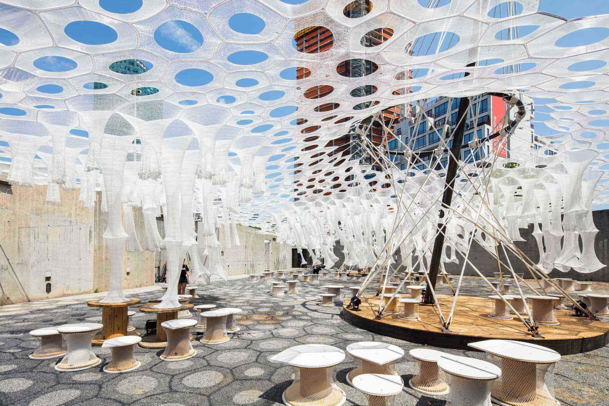 Installation Art 桌布 壁紙 — 一個未來感白色蜂巢天篷保護著陽光普照的城市庭院中的現代編織椅子。