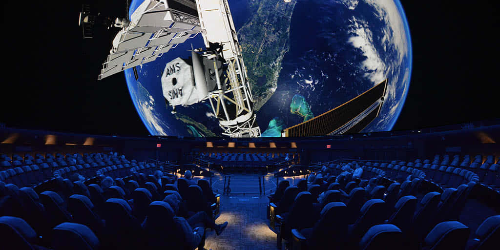 Planetarium 桌布 壁紙 — 一個未來風格的劇院，座位排列面向一個巨大的穹頂螢幕，顯示衛星在夜間繞地球運行。