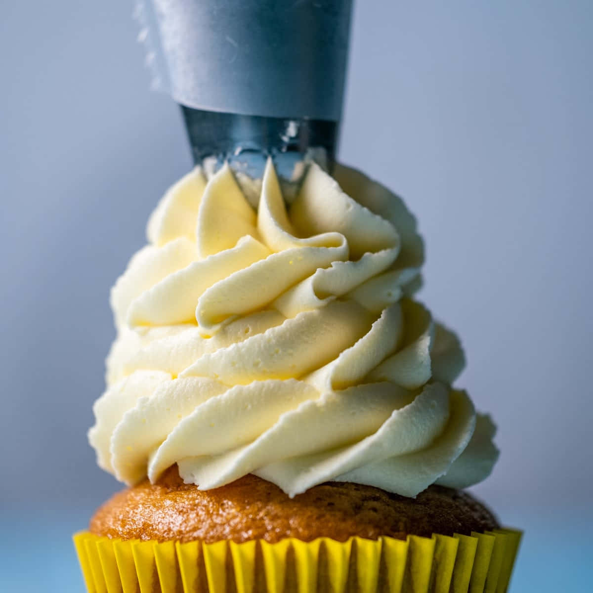 Buttercream 桌布 壁紙 — 一個杯子蛋糕在黃色襯紙中，正在被擠上奶油香草糖霜，背景是柔和藍色的特寫。