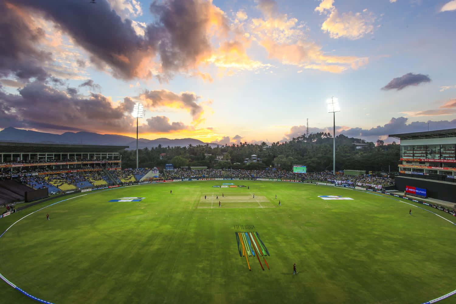 Cricket Ground 桌布 壁紙 — 一個板球場沐浴在金色的夕陽餘暉中，球員們在翠綠的草地上比賽，背景是連綿的山脈。