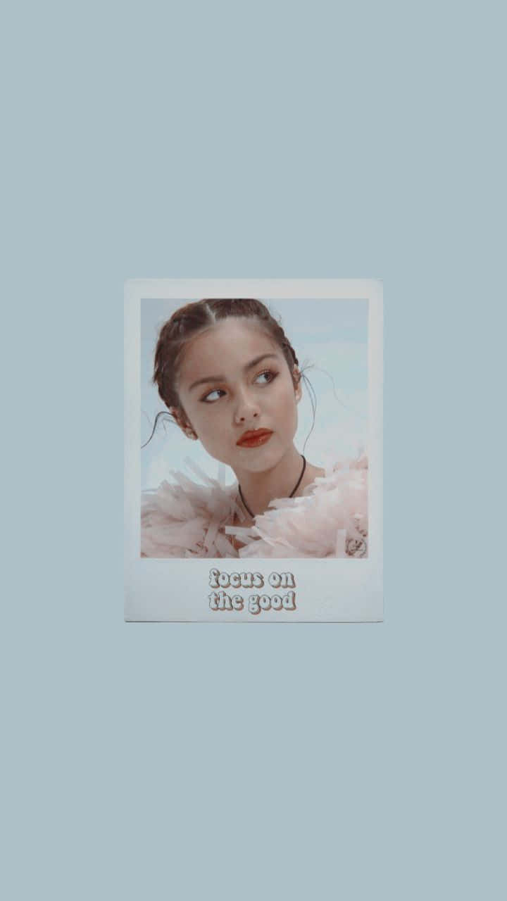 Olivia Rodrigo Aesthetic 桌布 壁紙 — 一個柔和的拍立得風格肖像，一個女人穿著毛茸茸的粉紅色領子，背景是淡藍色，上面寫著專注於美好事物。