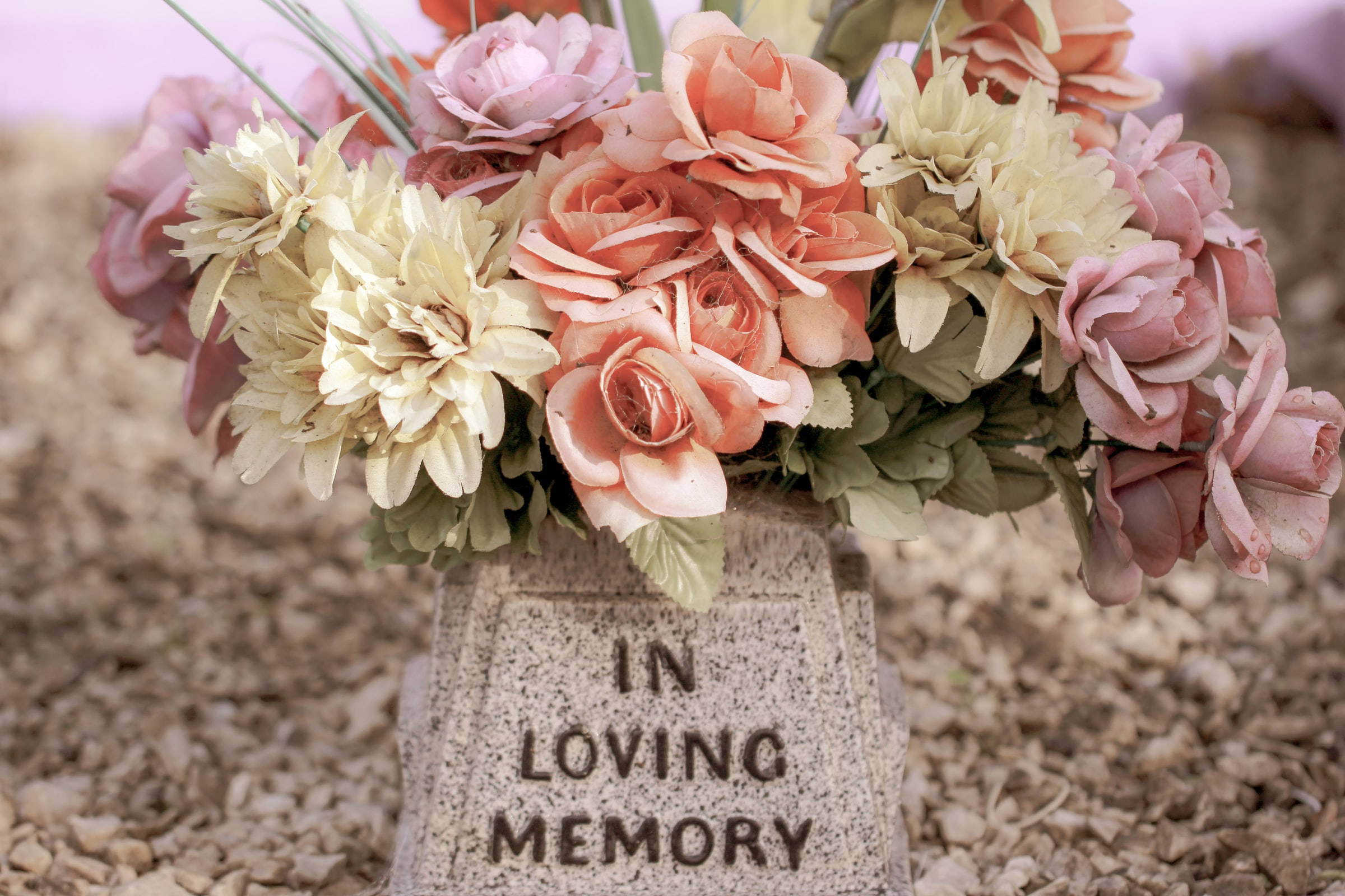 Remembrance 桌布 壁紙 — 一個柔和粉彩的花藝擺設放在刻有「IN LOVING MEMORY」的墓碑上，處於寧靜的墓園環境中。