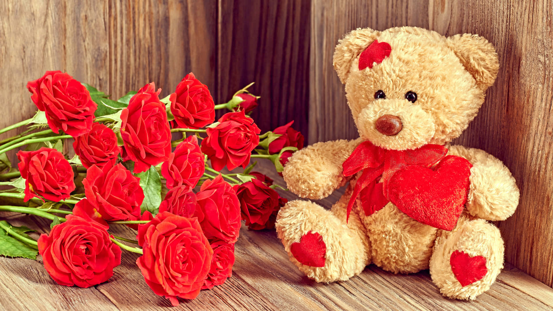 Cute Teddy 桌布 壁紙 — 一個柔軟的泰迪熊帶有紅色心形裝飾，坐在一個質樸的木質表面上，旁邊是一束鮮豔的紅玫瑰。