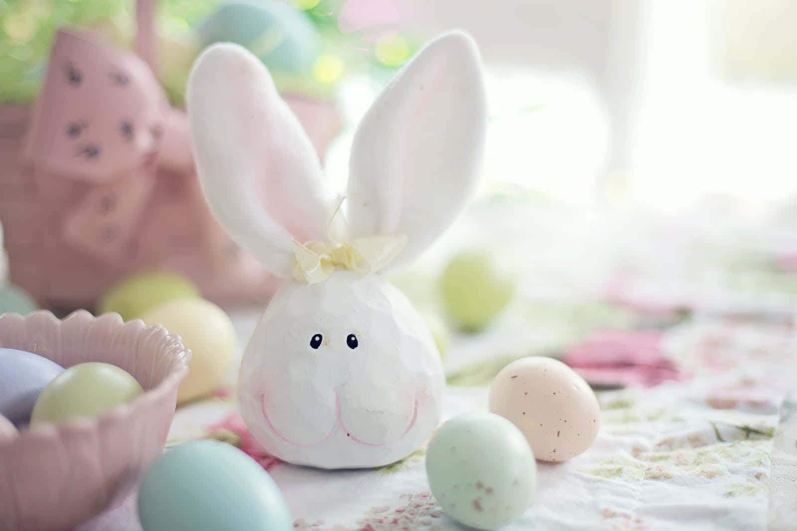 Easter Bunny 桌布 壁紙 — 一個柔軟的白色兔子公仔，繫著黃色蝴蝶結，坐在花桌布上的粉彩復活蛋之間。
