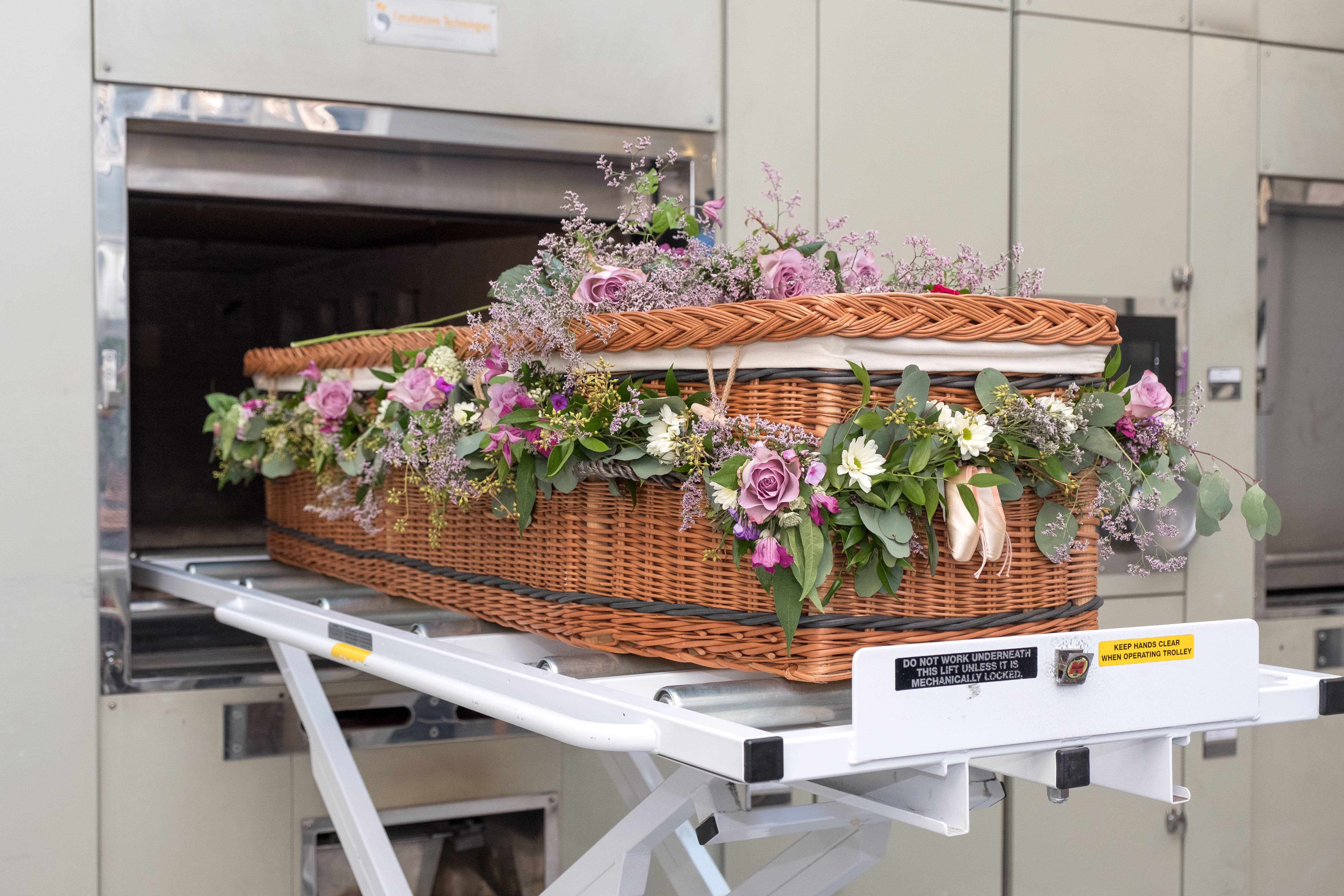 Funeral 桌布 壁紙 — 一個柳條棺材，飾有紫色玫瑰和綠色植物，正在白色推車上被移向不鏽鋼火化爐。