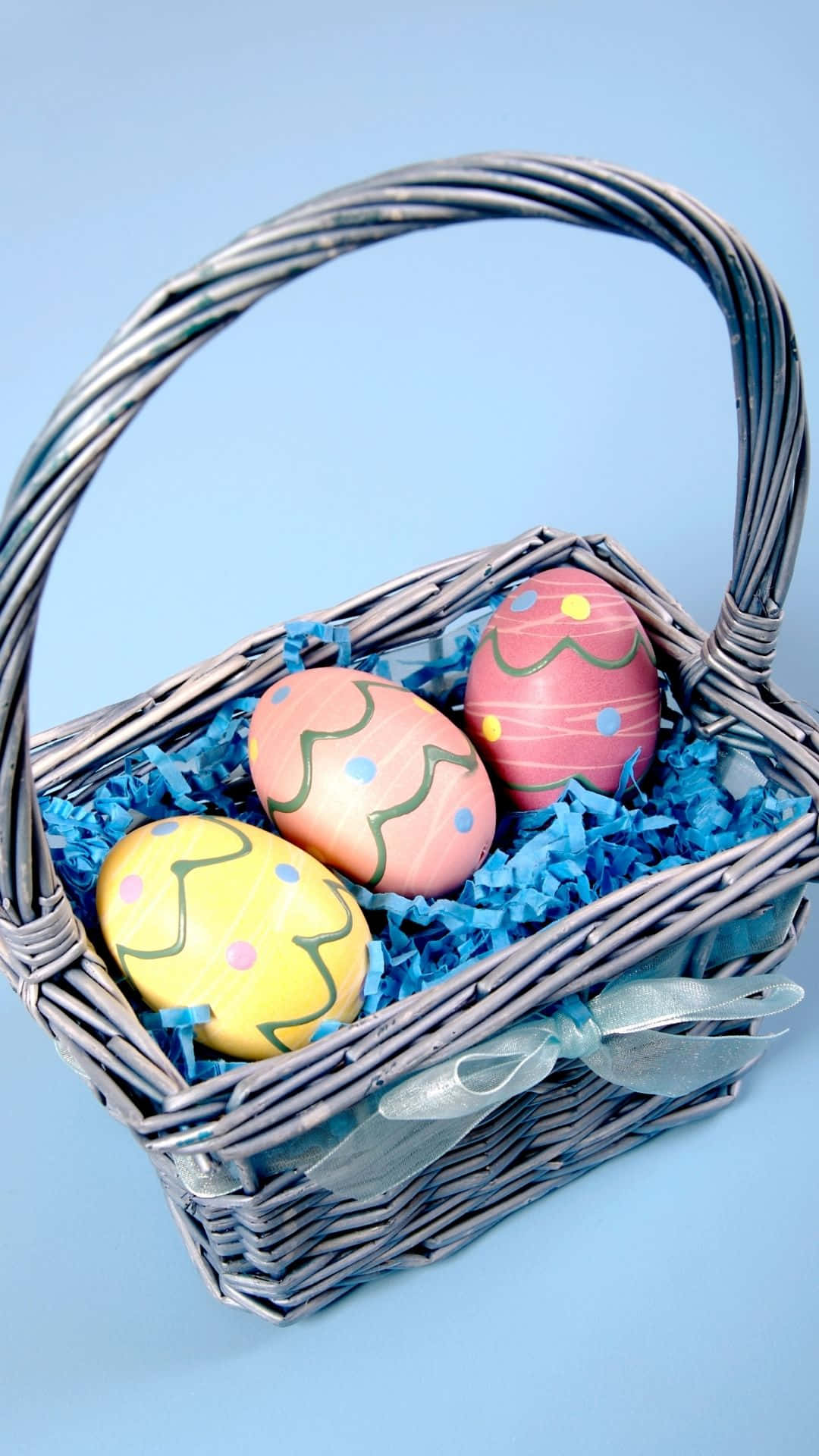Easter Basket 桌布 壁紙 — 一個柳條籃子，裡面裝滿了色彩繽紛的手繪復活節彩蛋，放在藍色碎紙中，襯著柔和的藍色背景。