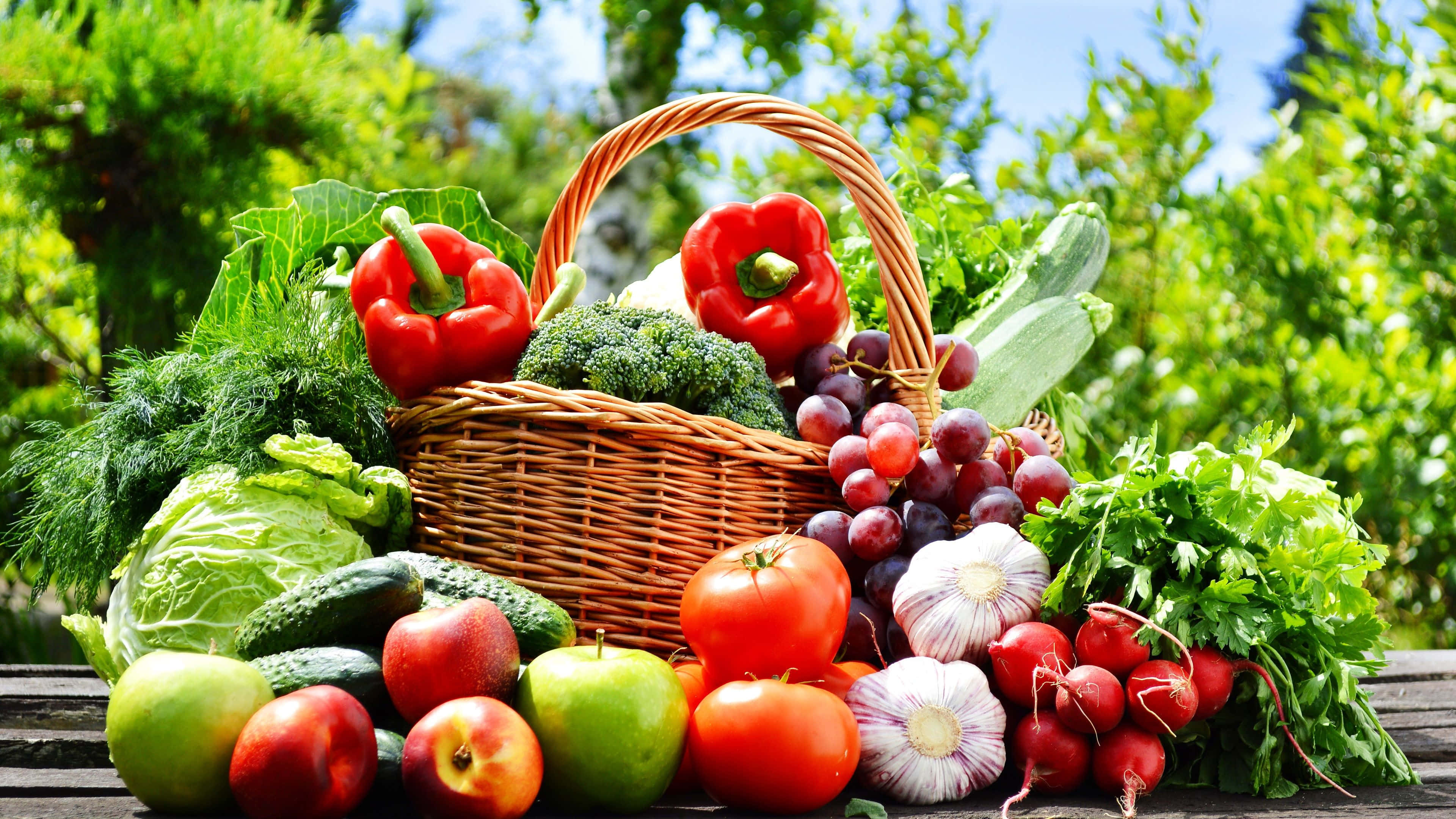 Fruits 桌布 壁紙 — 一個柳條籃子裝滿了新鮮的紅椒、番茄、葡萄和香草，放在木桌上，沐浴在明亮的陽光下。