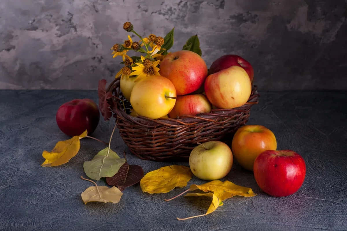 Winter Fruits 桌布 壁紙 — 一個柳條籃子裝滿了紅色和黃色的蘋果和向日葵，放在一個灰色表面上，周圍散落著秋天的落葉。