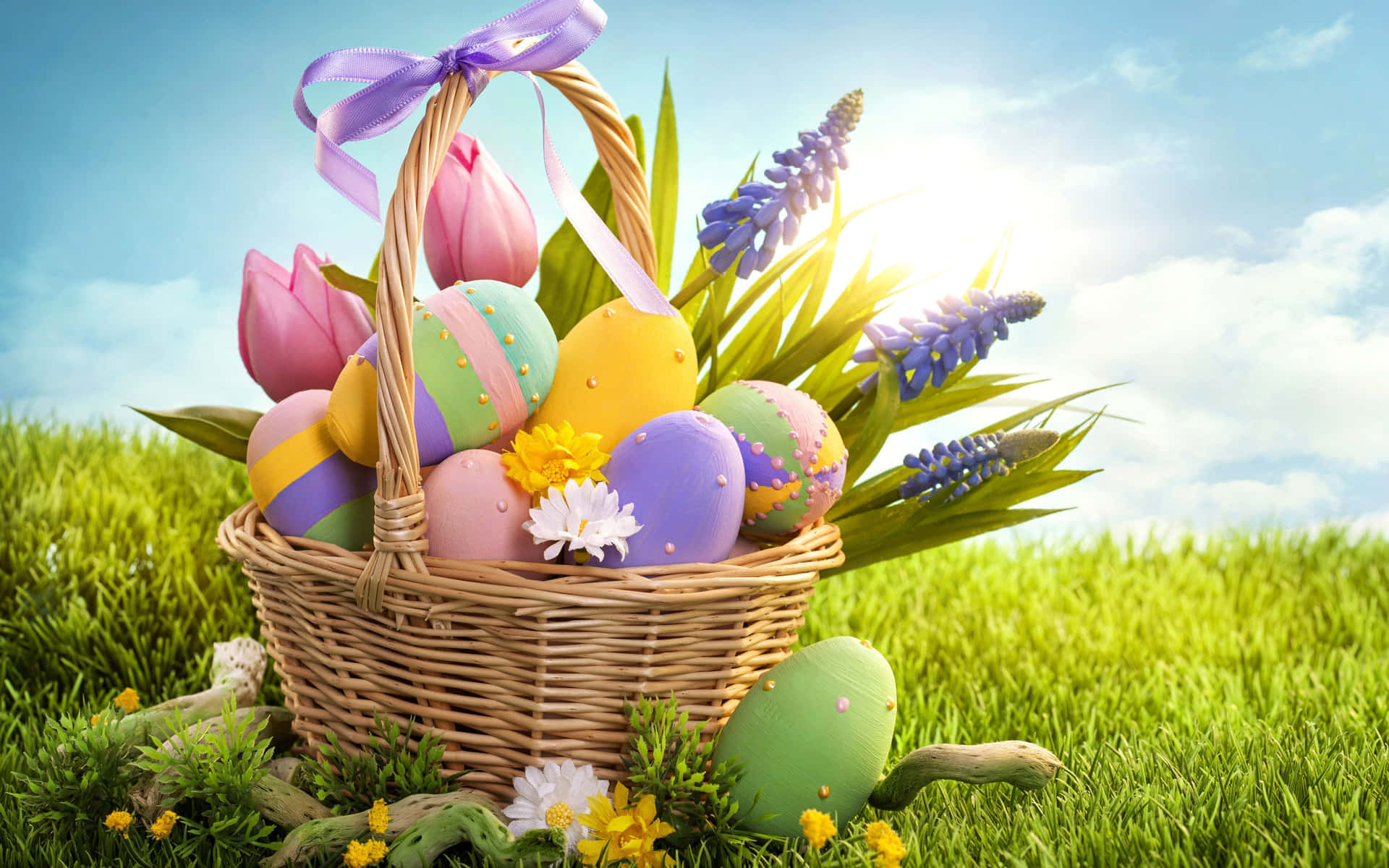 Easter 桌布 壁紙 — 一個柳條籃子裝滿了色彩繽紛的復活節彩蛋和春天的花朵，放在充滿活力的綠色草地上，頭頂是明亮的藍天。