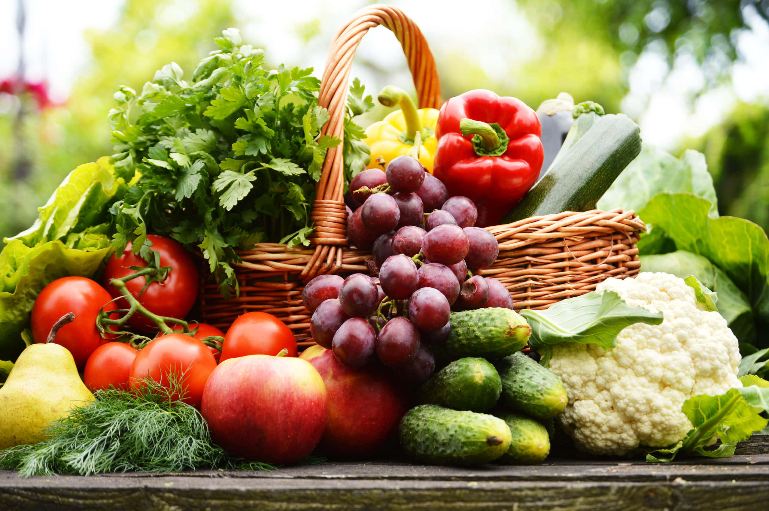 Fruits 桌布 壁紙 — 一個柳條籃子裝滿新鮮農產品，包括葡萄、番茄和花椰菜，背景是模糊的綠色花園。