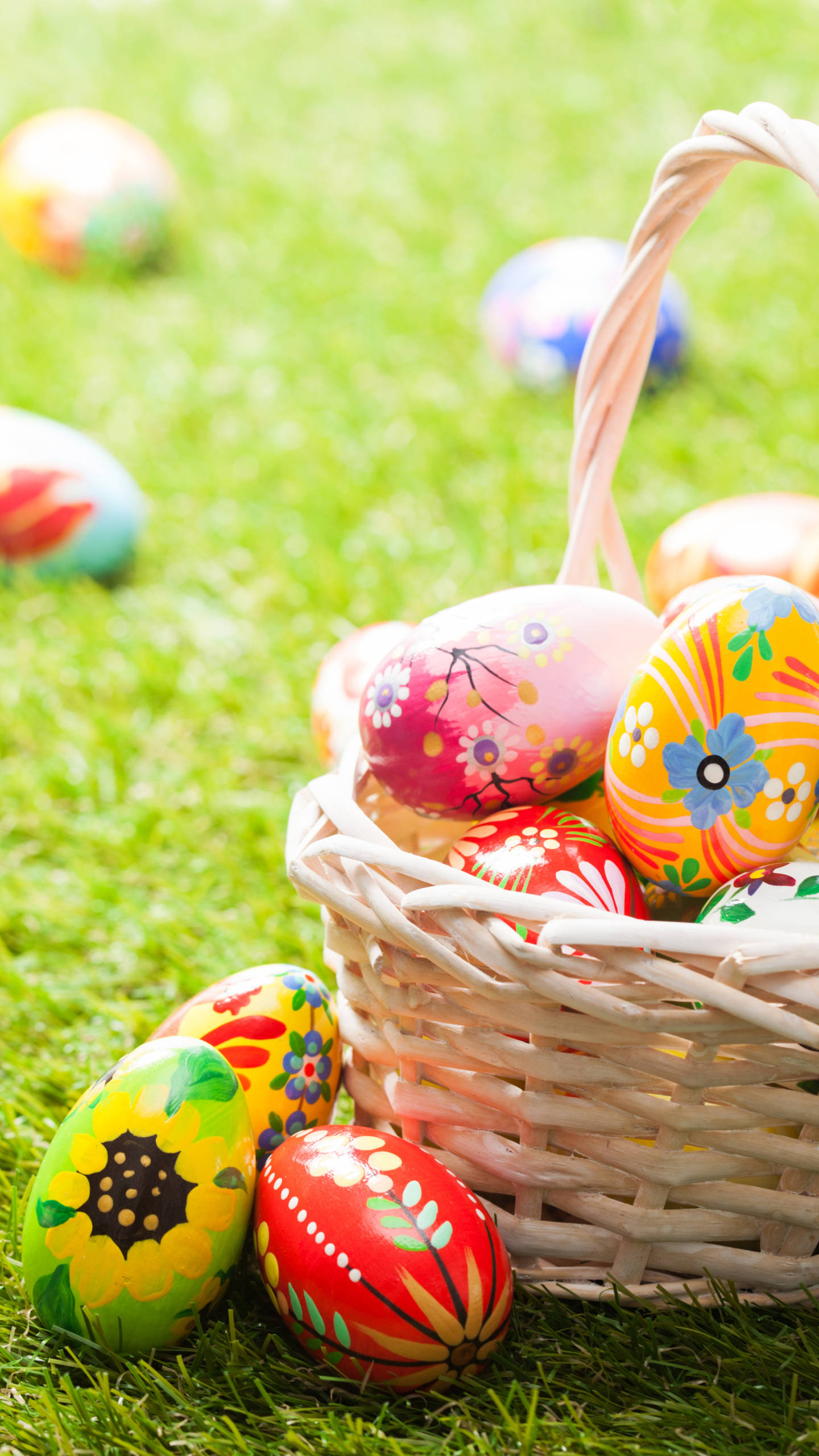 Easter Phone 桌布 壁紙 — 一個柳條籃子裝滿色彩鮮豔的復活節彩蛋，放在茂密的綠草地上，附近還有更多散落的彩蛋。