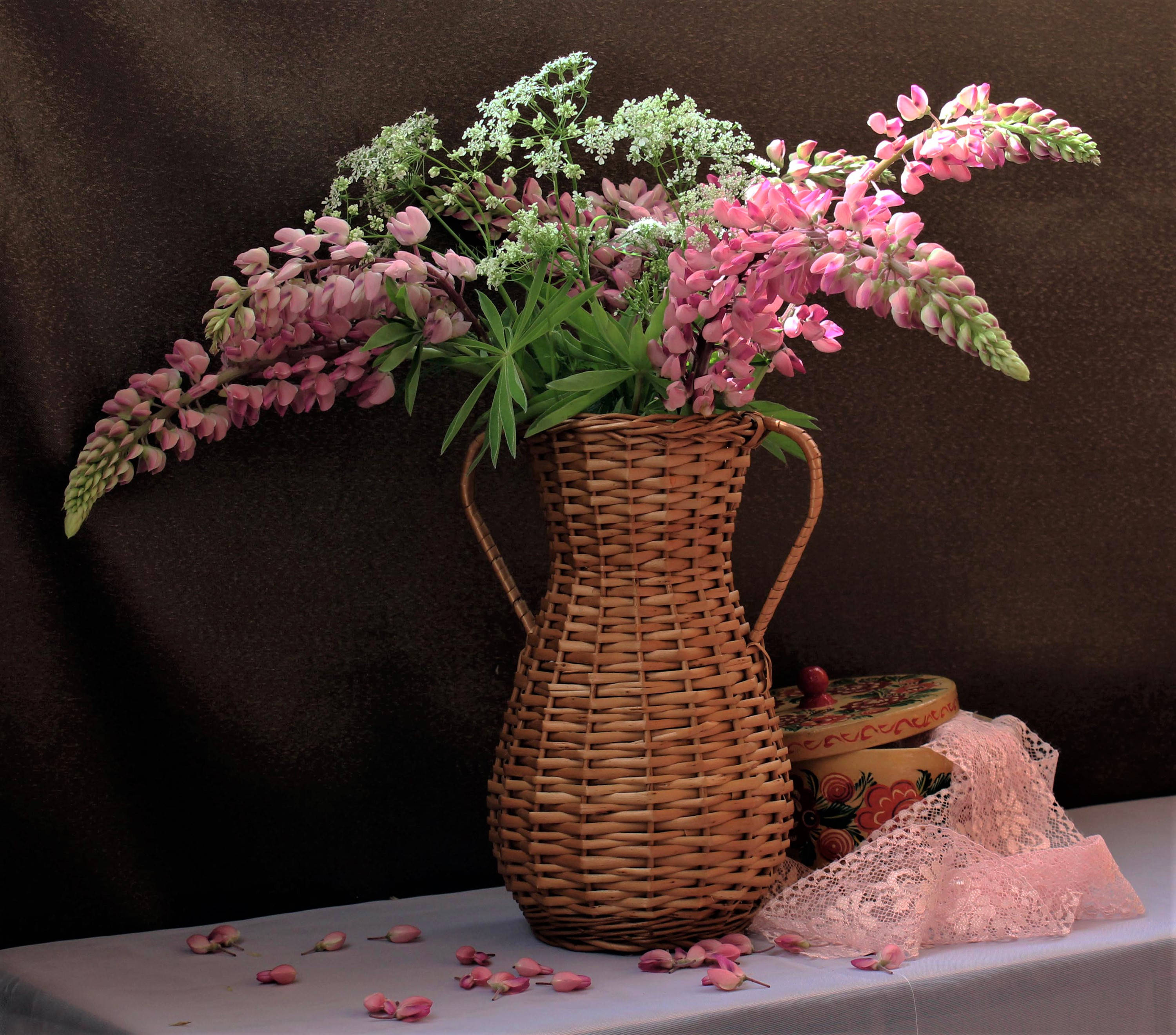 Bouquet 桌布 壁紙 — 一個柳條花瓶裡插滿了粉紅色的羽扇豆和滿天星，放在一塊白布上，旁邊有蕾絲和一個花盒，背景是深色的。