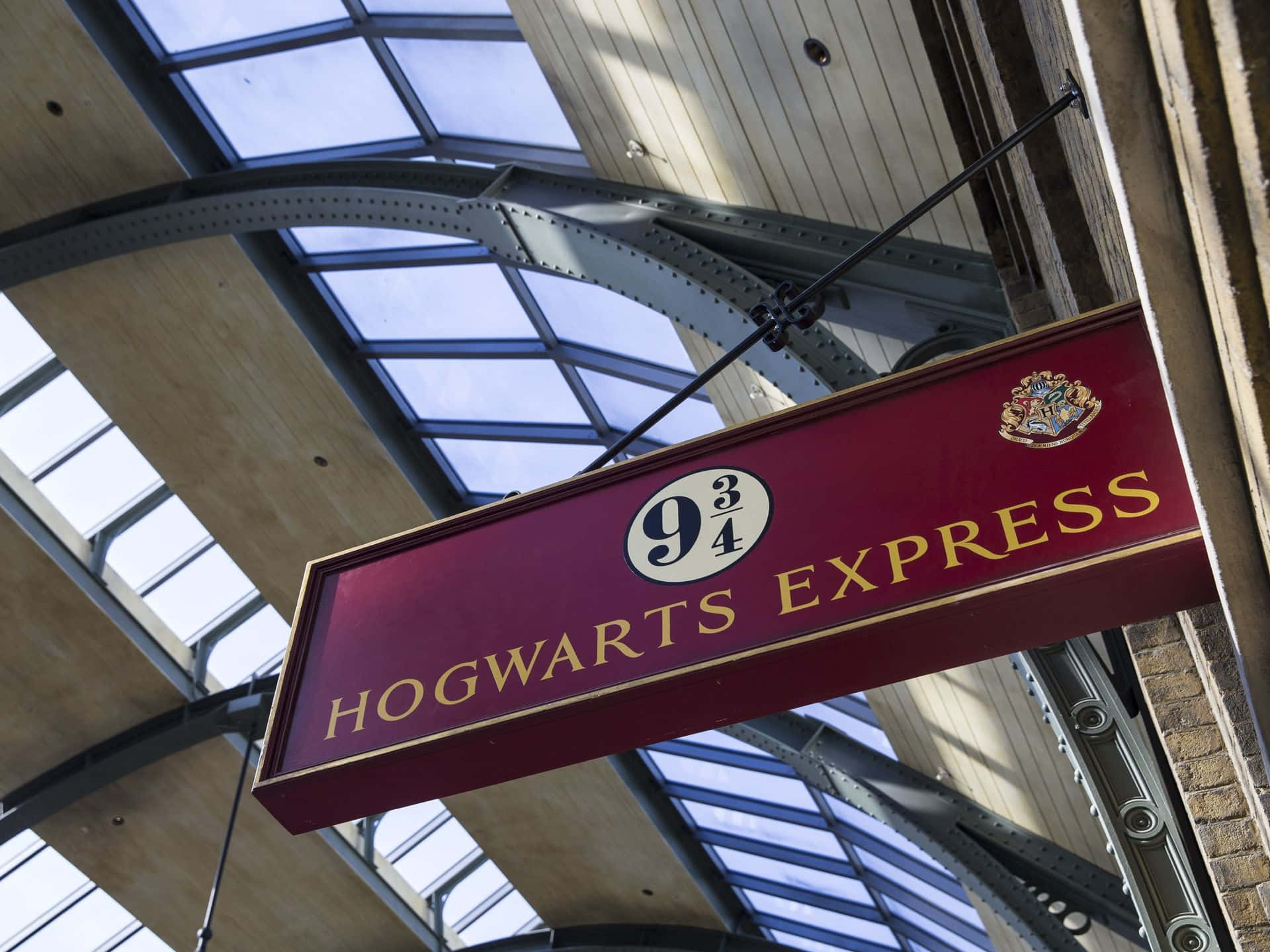 Hogwarts Express 桌布 壁紙 — 一個栗色的霍格華茲特快列車標誌懸掛在玻璃和鋼結構的車站屋頂下，沐浴在日光中。