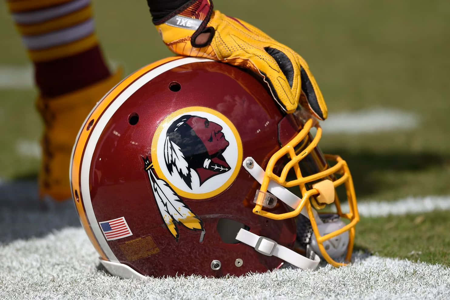 Washington Football 桌布 壁紙 — 一個栗色華盛頓紅皮隊美式足球頭盔，帶有黃色面罩，放在草地上，一隻戴手套的手觸碰其頂部。