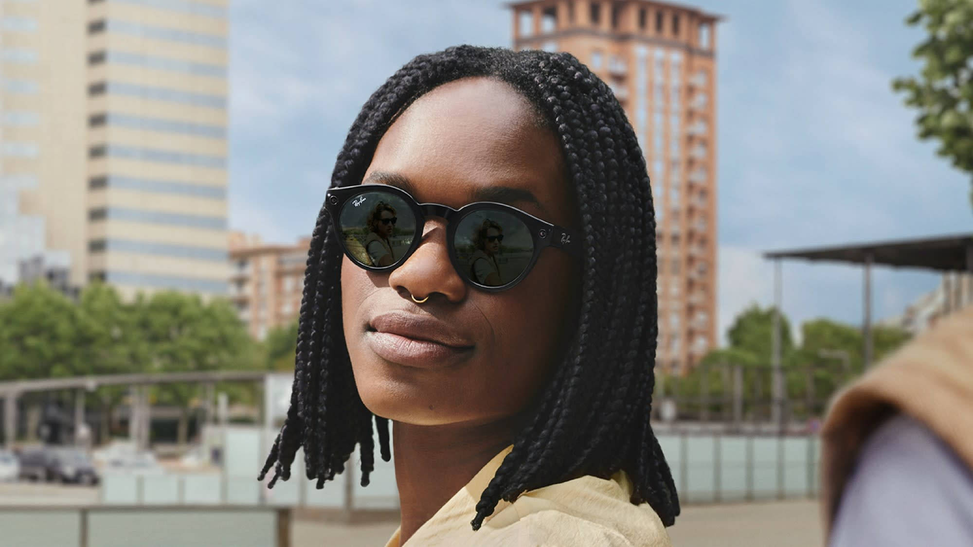 Sunglasses 桌布 壁紙 — 一個梳著辮子、戴著太陽眼鏡的人自信地站在陽光普照的城市廣場上，背景是現代建築和綠樹。