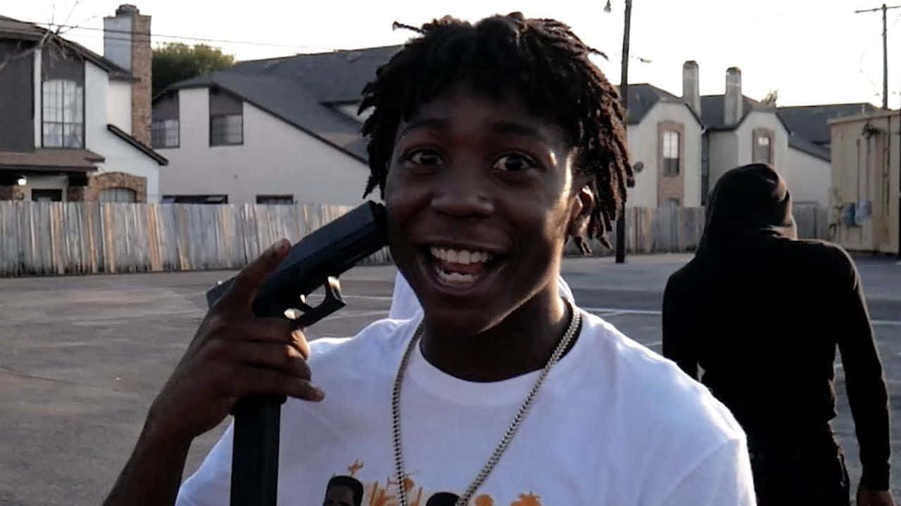 Rappers 桌布 壁紙 — 一個梳著辮子的年輕人在黃昏的郊區小巷中，咧嘴笑著將槍指著自己的頭。