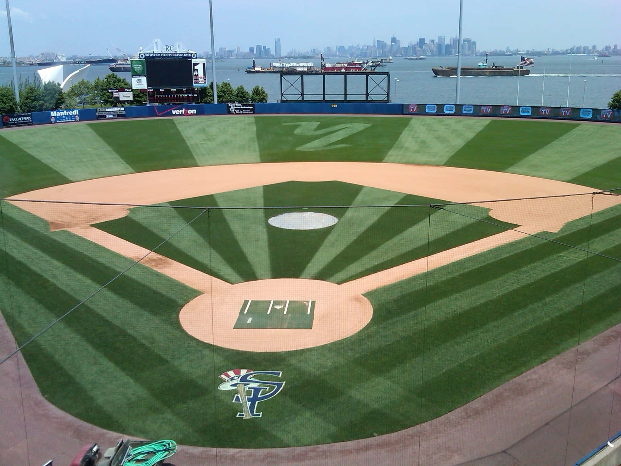 Baseball Field 桌布 壁紙 — 一個棒球場的鳥瞰圖，有條紋綠色草地和棕色泥土內野，俯瞰城市天際線和水面。