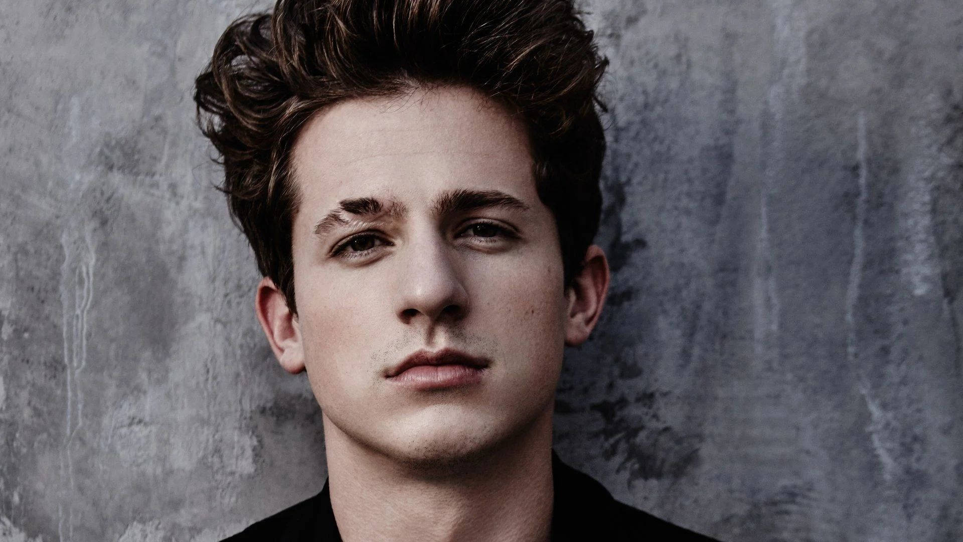 Charlie Puth 桌布 壁紙 — 一個棕色亂髮的年輕男子，在紋理灰色混凝土牆前直視鏡頭。