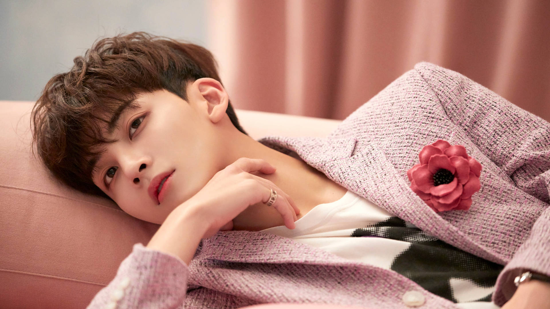 yoon jeonghan 桌布 壁紙 — 一個棕色亂髮的年輕男子將頭靠在粉紅色沙發上，身穿帶有紋理的粉紅色西裝外套，飾有紅色花朵別針。