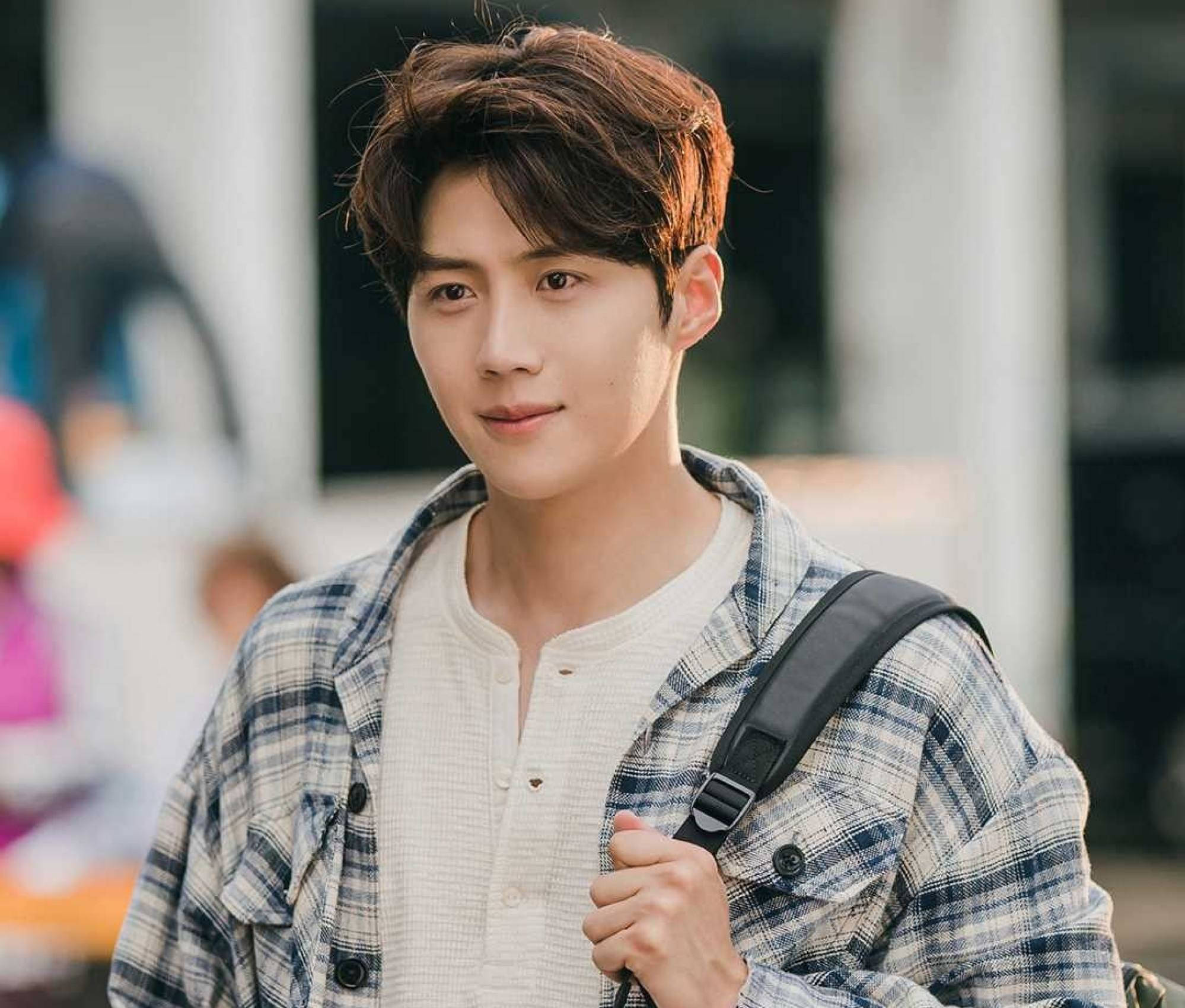 Kim Seon Ho 桌布 壁紙 — 一個棕色亂髮的年輕男子穿著格紋襯衫，背著背包，溫柔地微笑。