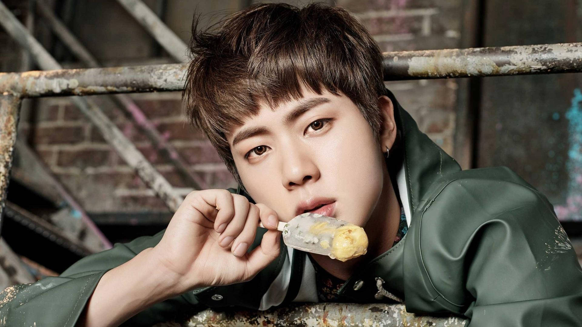 kim seok jin 桌布 壁紙 — 一個棕色亂髮的年輕男子穿著綠色外套，在粗糙的磚牆和金屬背景下咬著融化的冰淇淋甜筒。