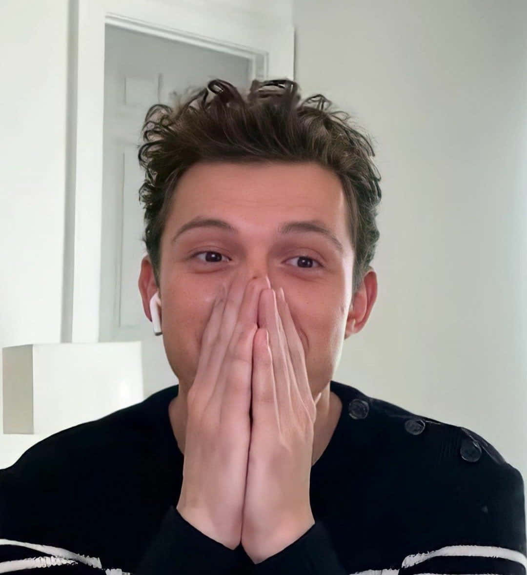 Tom Holland Aesthetic 桌布 壁紙 — 一個棕色亂髮的年輕男子雙手捂住嘴巴，戴著白色耳塞和黑色毛衣。