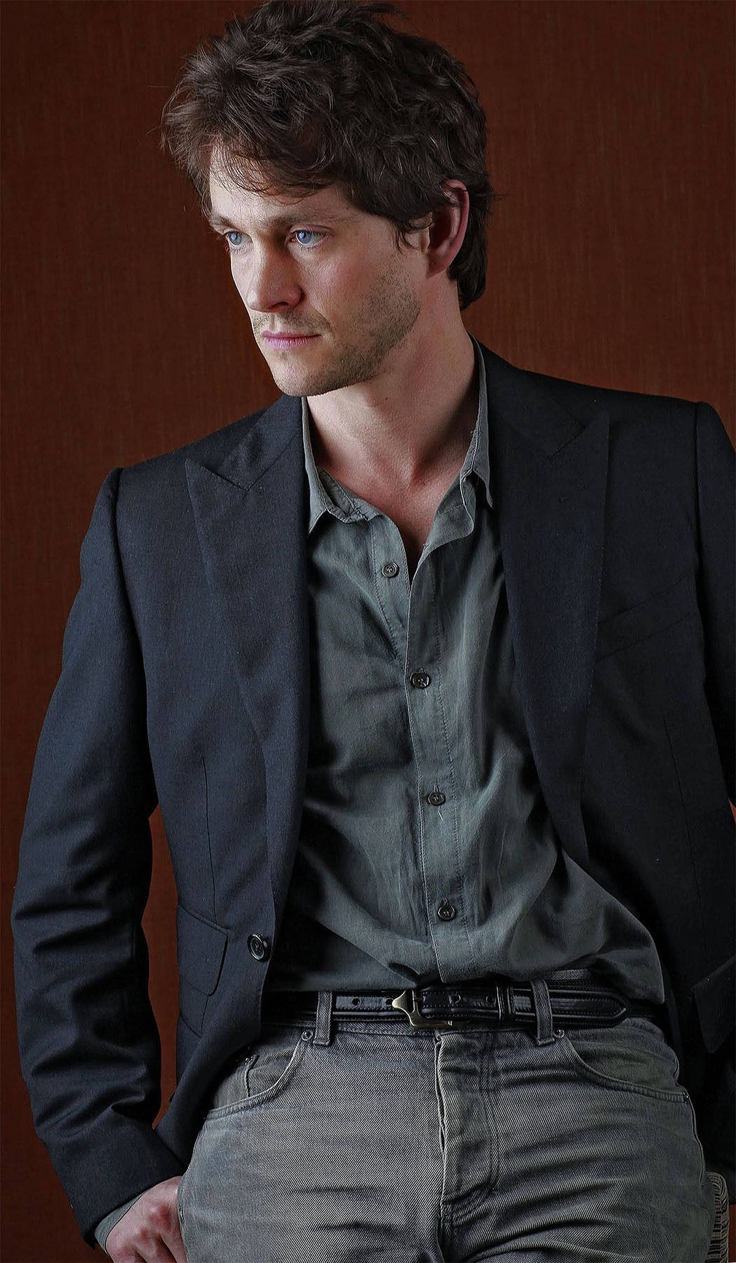 Hugh Dancy 桌布 壁紙 — 一個棕色亂髮藍眼的男人，穿著深色西裝外套，內搭灰色襯衫和牛仔褲，背景是溫暖的木質背景。