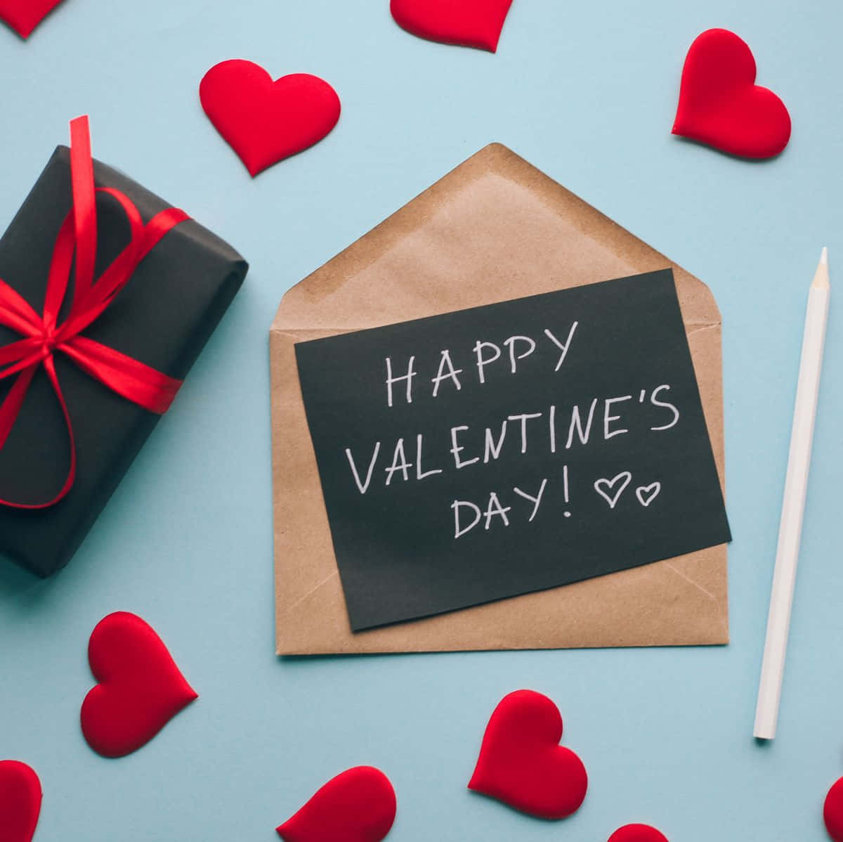 愛心 桌布 壁紙 — 一個棕色信封的平鋪構圖，附有寫著 'Happy Valentine's Day' 的黑板便條，旁邊是一個繫著紅絲帶的黑色禮物盒，淺藍色表面上散落著紅色心形。