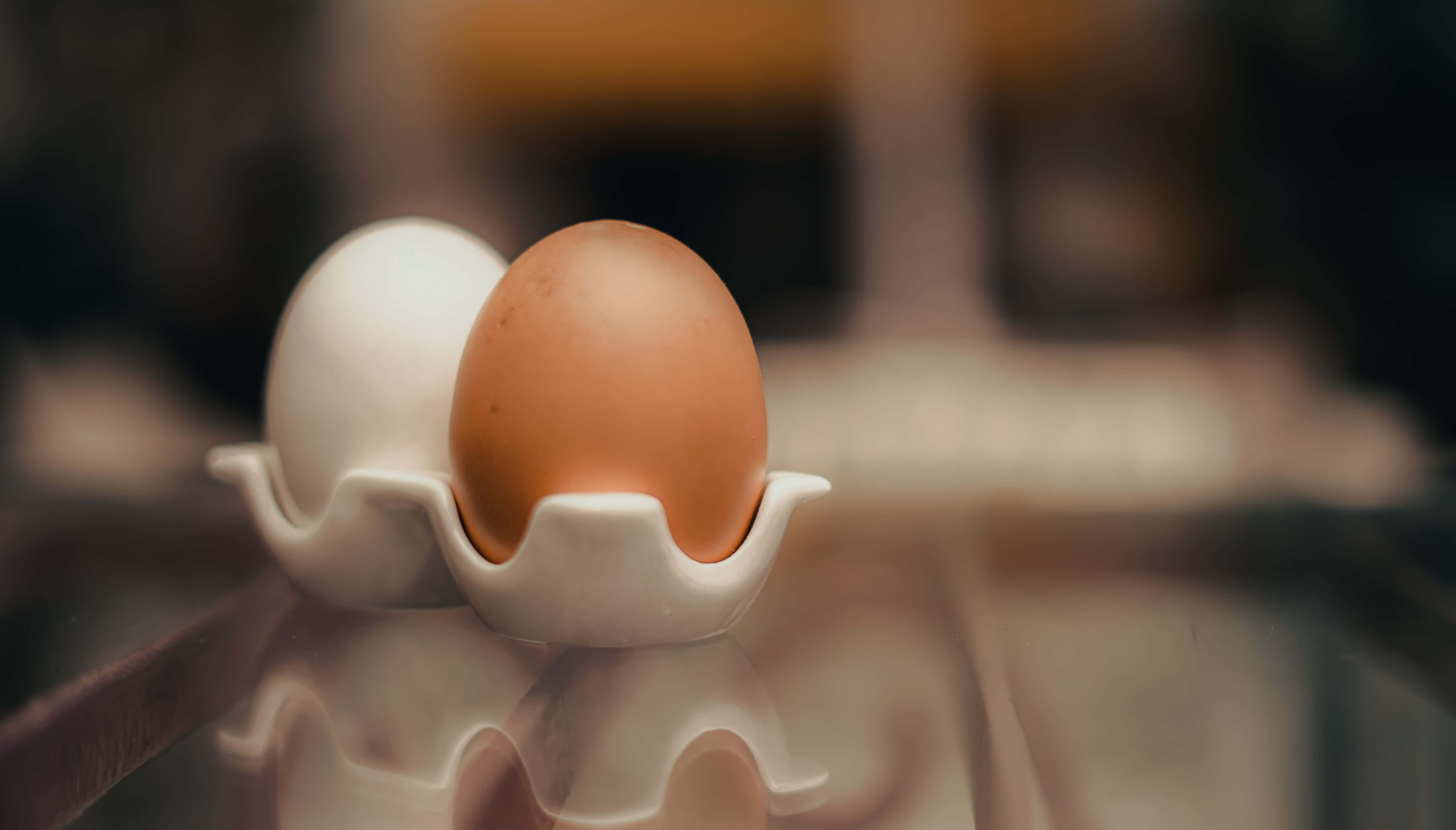Brown Eggs 桌布 壁紙 — 一個棕色和白色雞蛋的特寫，放在白色陶瓷蛋架上，置於反射表面，帶柔和溫暖的燈光。