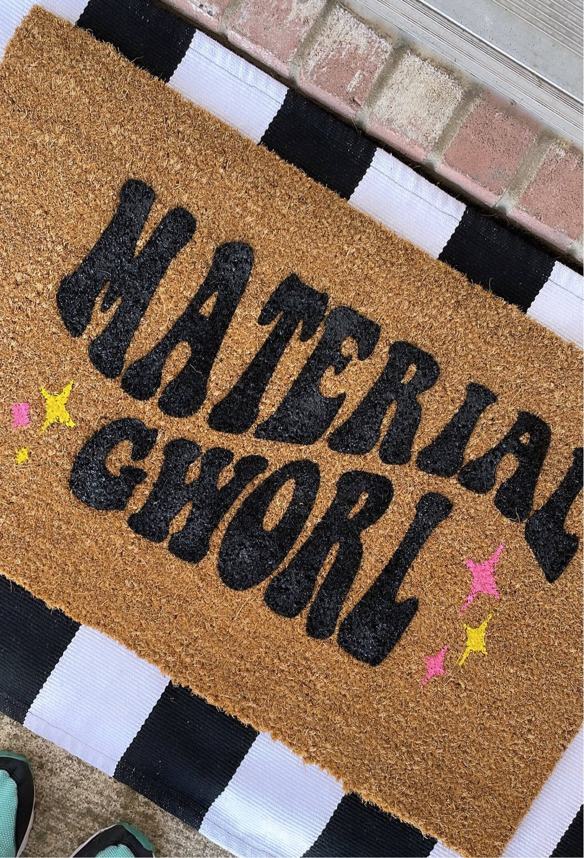 Interior Design 桌布 壁紙 — 一個棕色椰殼纖維門墊，上面有粗體黑色閃粉文字寫著「MATERIAL GHWORL」和彩色星星裝飾，放在磚門口的黑白條紋地毯上。