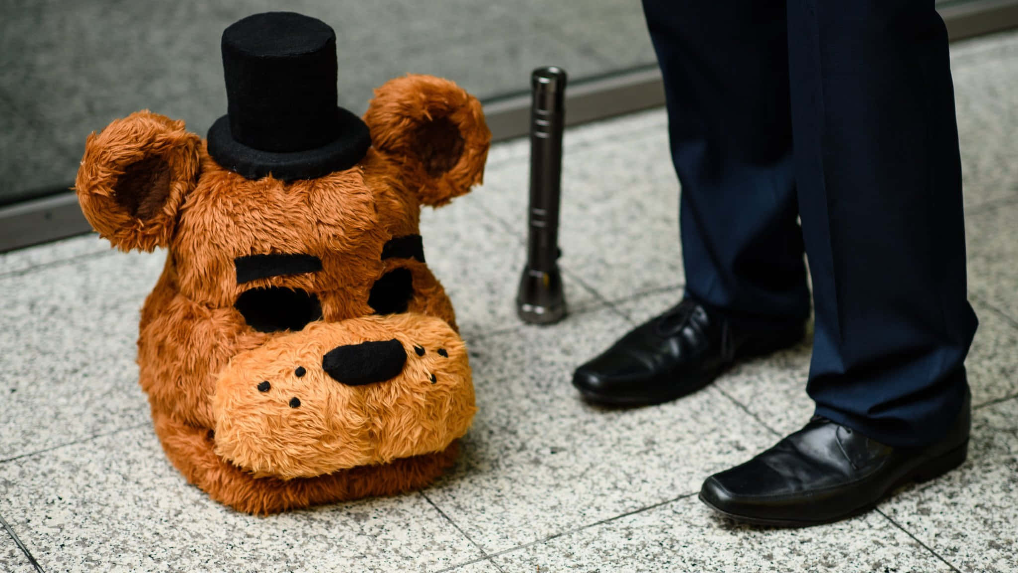 Five Nights At Freddys 桌布 壁紙 — 一個棕色毛茸茸的Freddy Fazbear頭戴黑色高帽，坐在磁磚地上，旁邊有一對人的腿和一個護柱。
