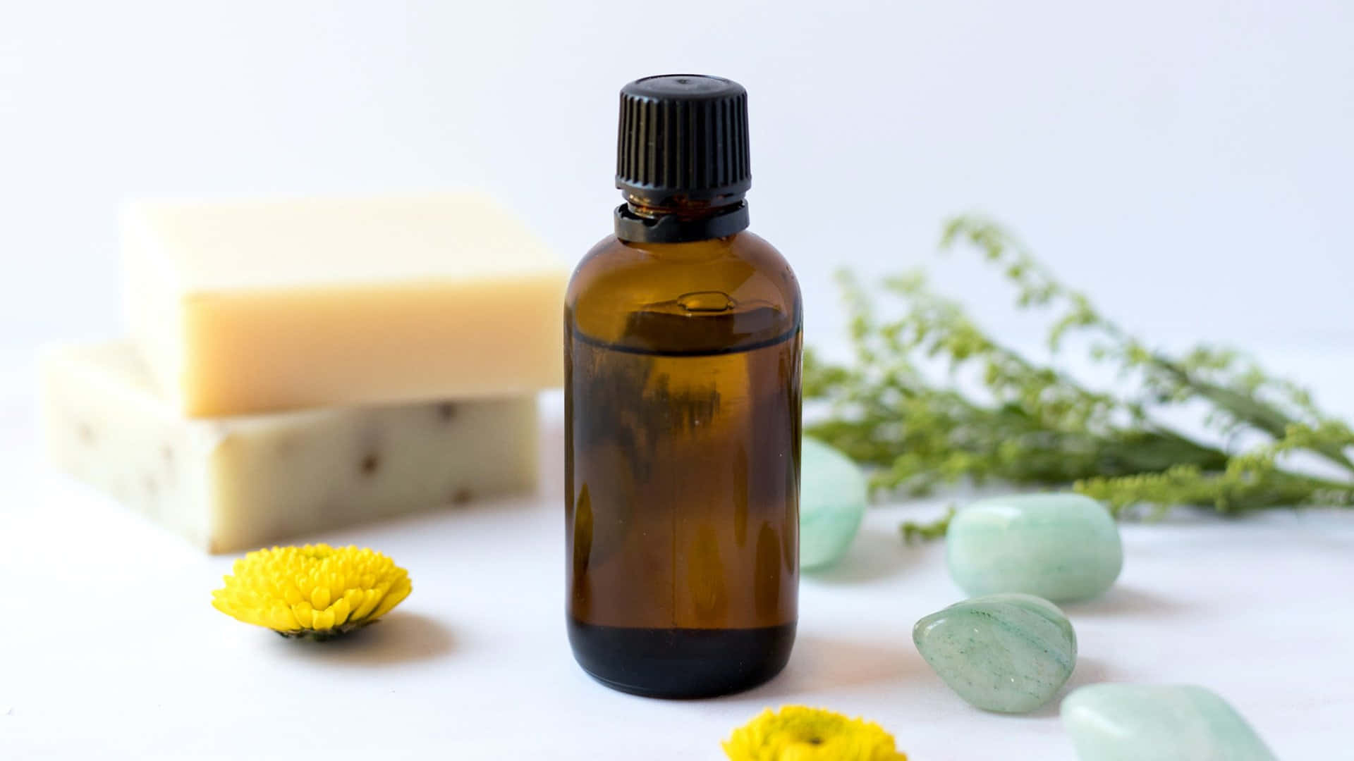 Aromatherapy 桌布 壁紙 — 一個棕色玻璃精油瓶居中放置，旁邊有肥皂塊、綠色石頭和黃色花朵，置於明亮的白色表面上。