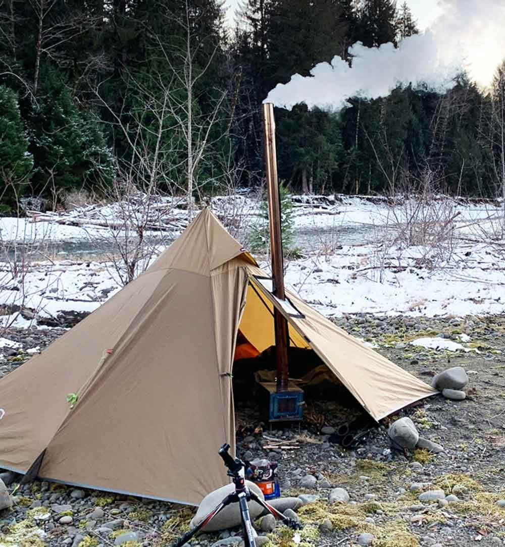 Winter Camping 桌布 壁紙 — 一個棕色的帳篷，帶有柴火爐煙囪冒出煙霧，矗立在白雪覆蓋的河岸旁，周圍是森林冬季景觀。