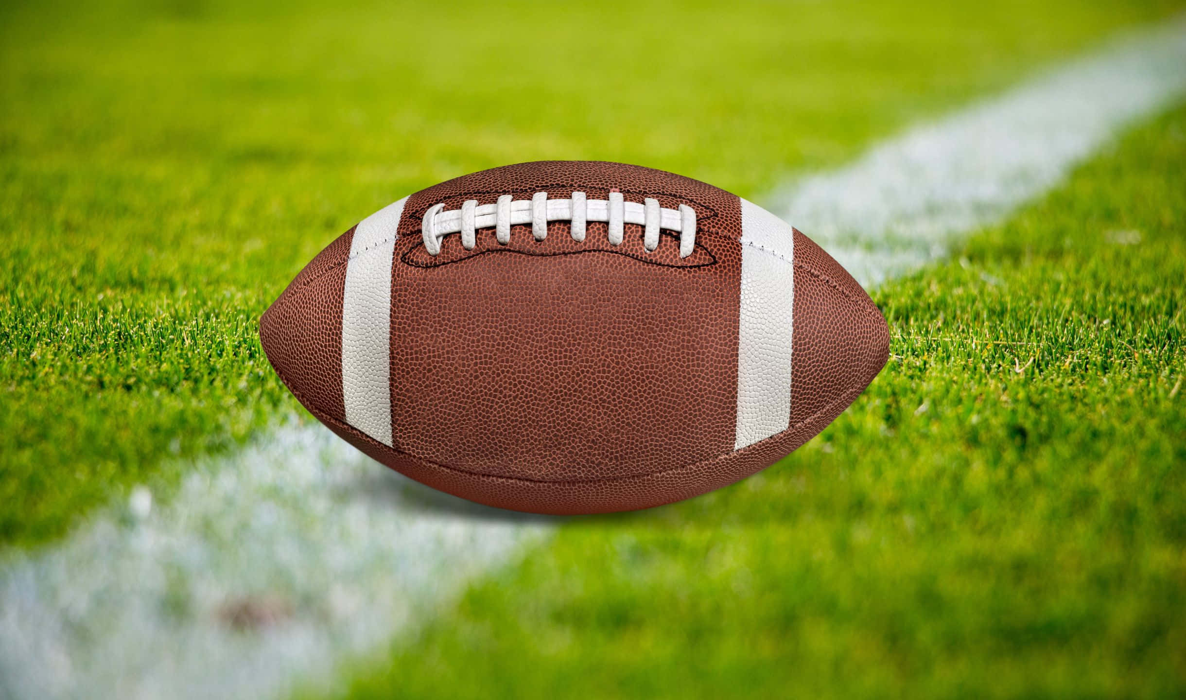 2440x1440 Football 桌布 壁紙 — 一個棕色皮革美式足球，帶有白色鞋帶和條紋，放在充滿活力的綠色草地上，旁邊是一條白色球場線。