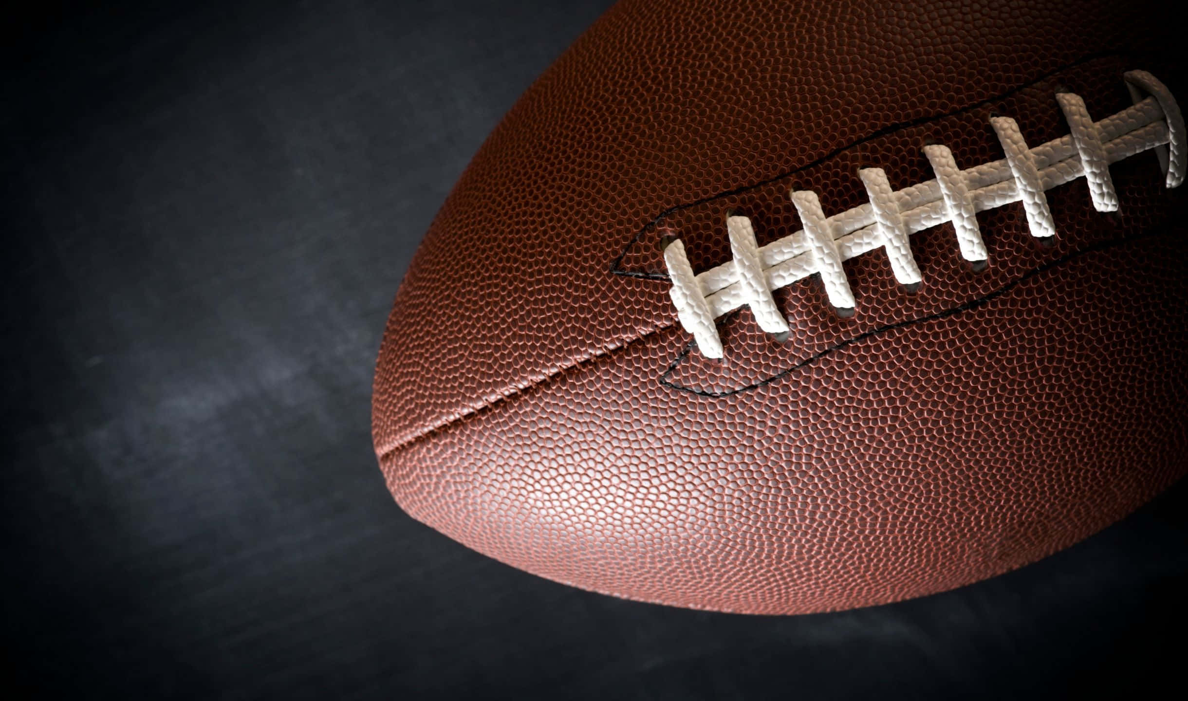 2440x1440 Football 桌布 壁紙 — 一個棕色皮革美式足球的特寫，帶有白色鞋帶，背景為深色紋理。