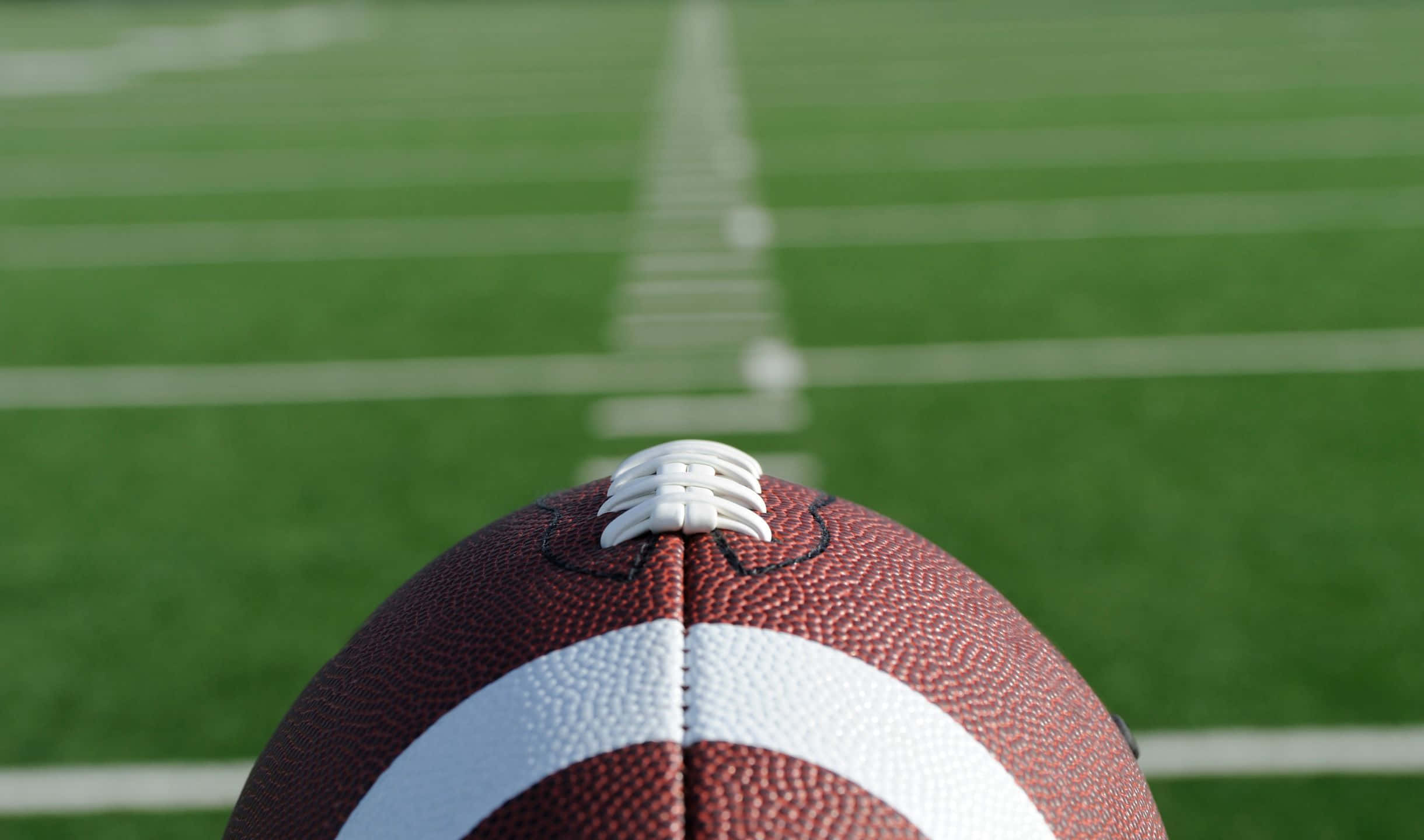 2440x1440 Football 桌布 壁紙 — 一個棕色皮革美式足球的特寫，帶有白色鞋帶，放置在充滿活力的綠色球場上，球場上有清晰的白色碼線。