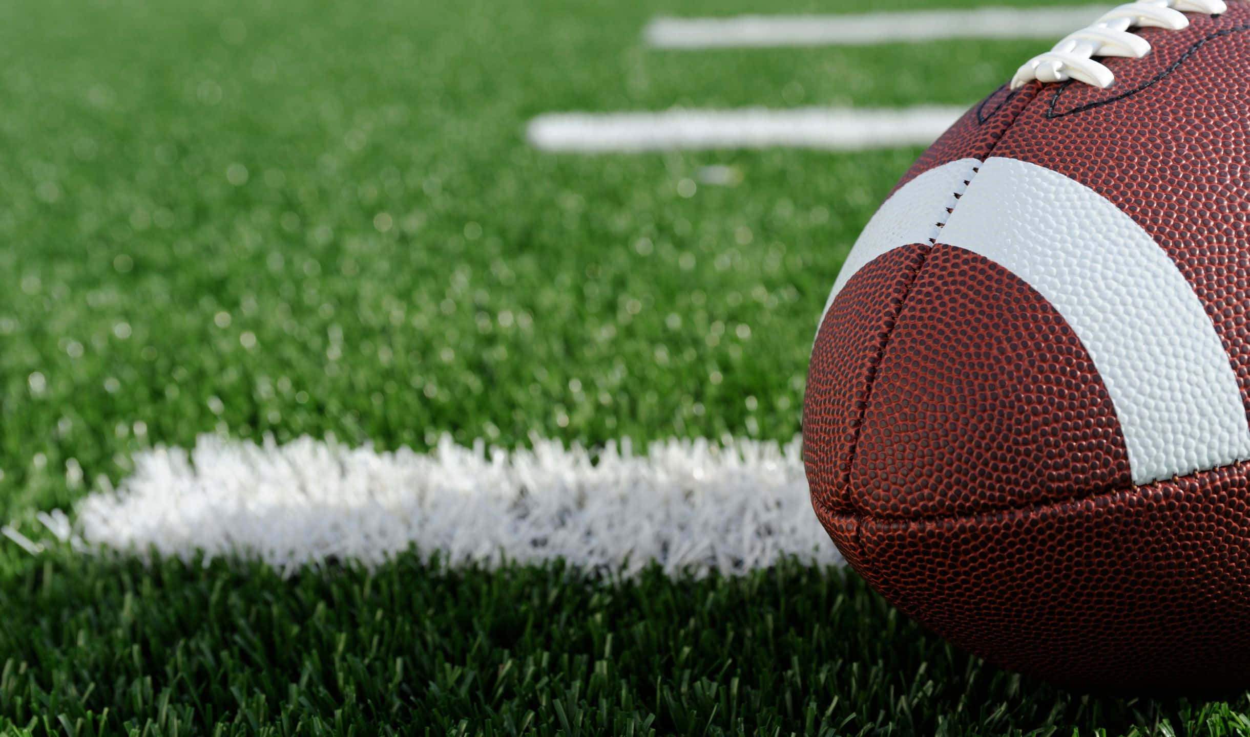 2440x1440 Football 桌布 壁紙 — 一個棕色皮革美式足球的特寫，放在充滿活力的綠色草地上，旁邊是一條清晰的白色碼線。